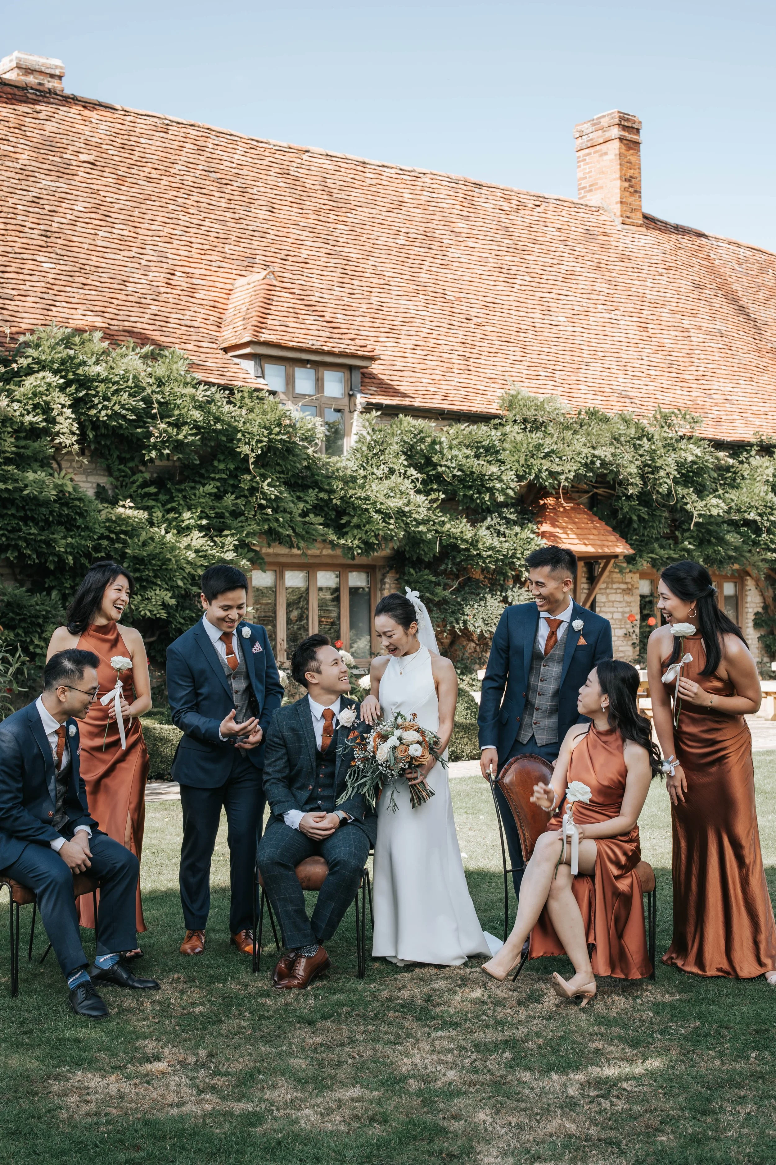 A wedding party outdoors in front of a house with a red-tiled roof. The group includes a bride, groom, and six attendants dressed in formal attire, laughing and engaged in conversation.