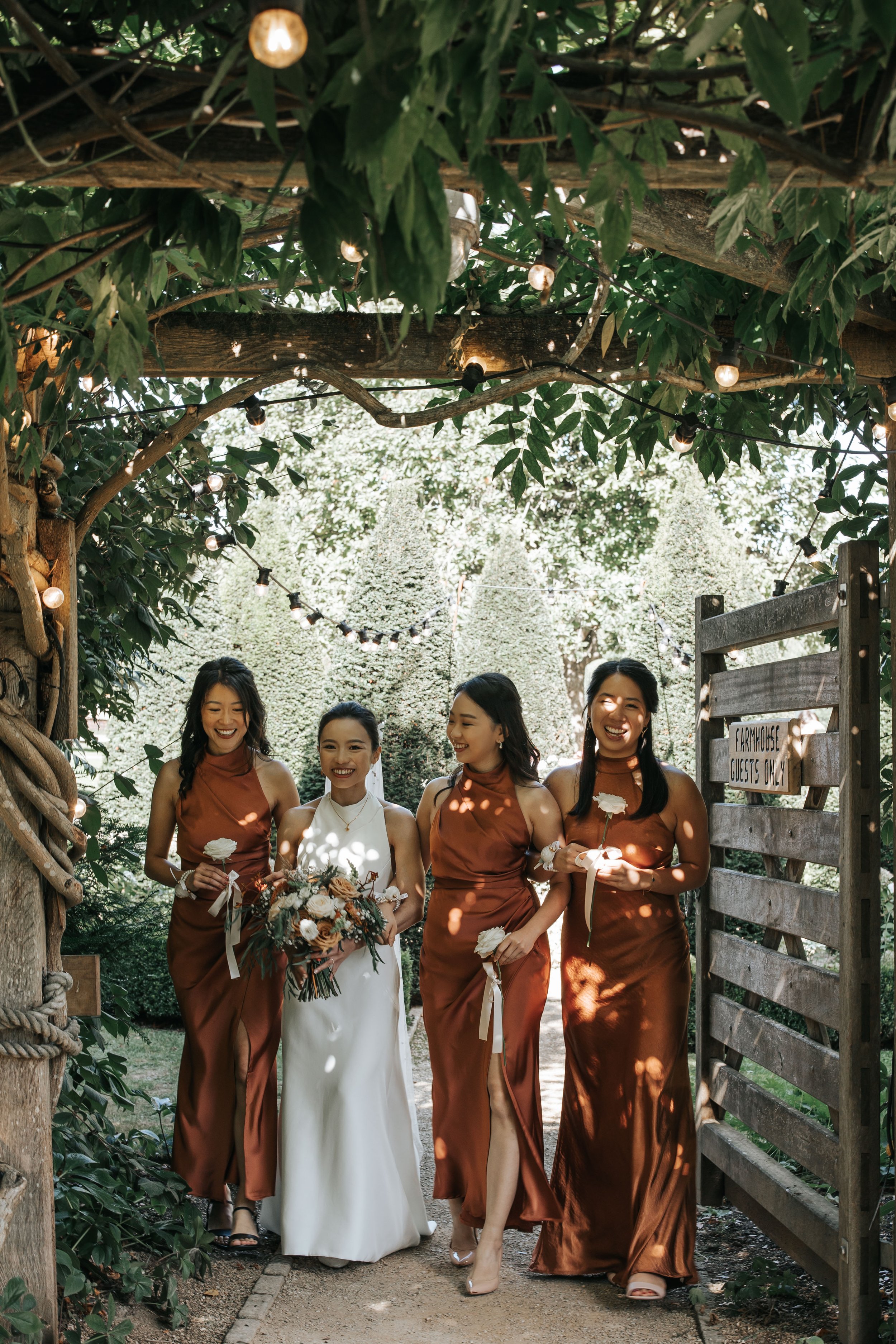 Four women, one in a white wedding dress and three in matching rust-colored bridesmaid dresses, walking through a garden archway decorated with string lights, smiling and holding flowers.