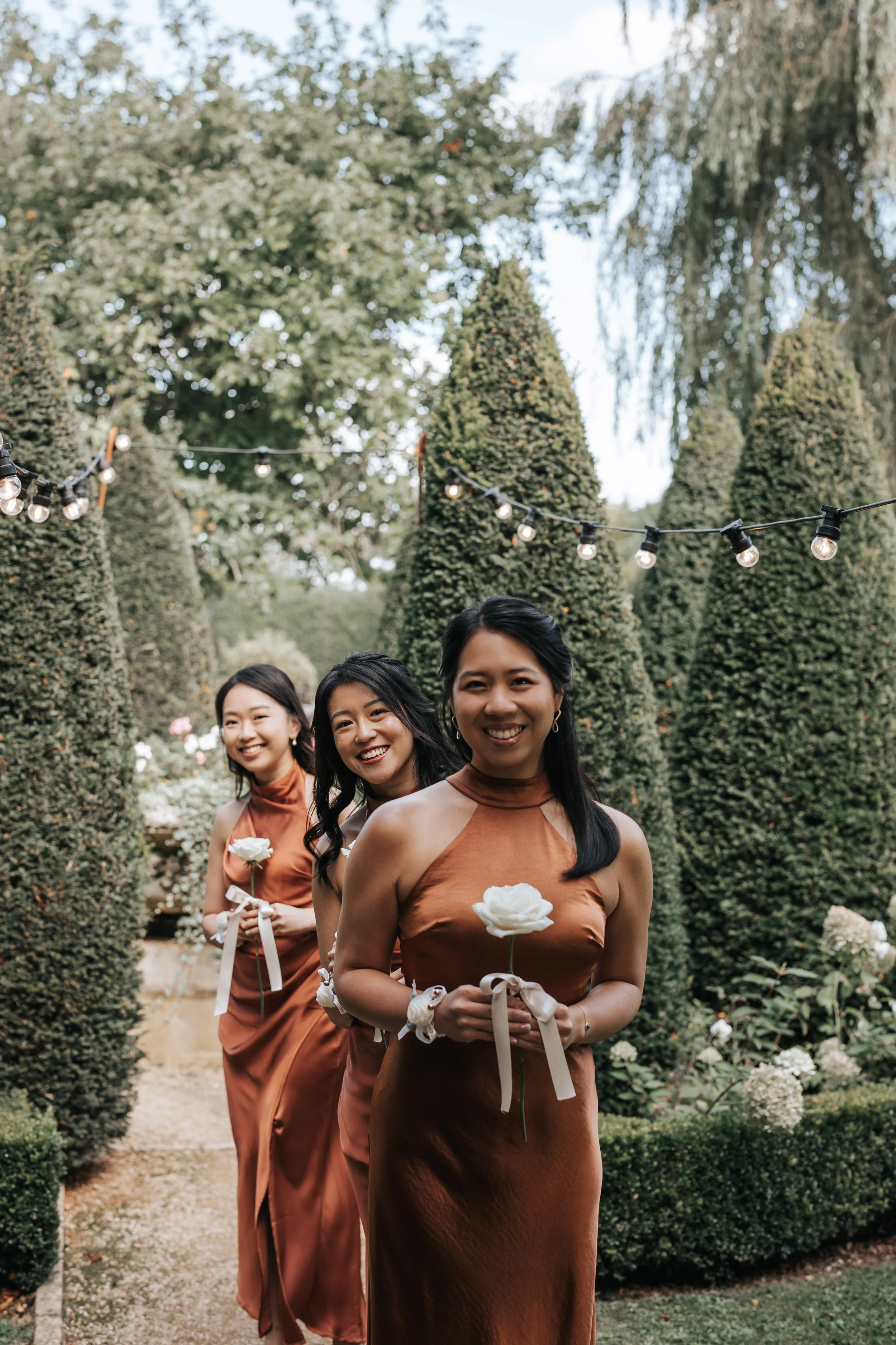 Three women dressed in matching rust-colored dresses standing in a garden with tall, shaped shrubs, holding white rose bouquets, smiling, with string lights overhead.