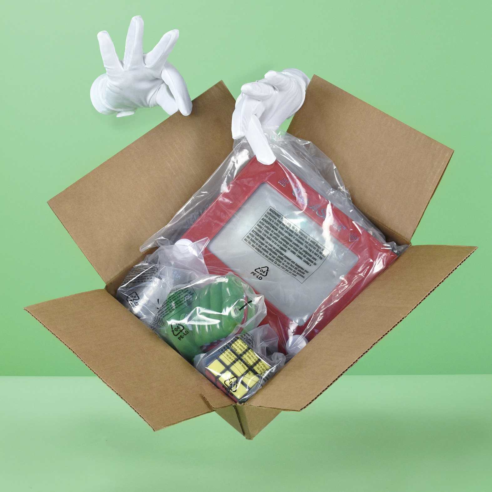 Open cardboard box with packing materials, including a wrapped item with a red frame, a Rubik's cube, a green object, and other items, on a green background.