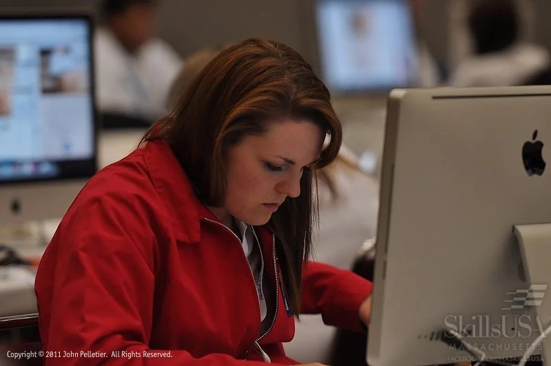 A woman with red hair wearing a red jacket, concentrating while working on a desktop computer in a classroom or office setting.