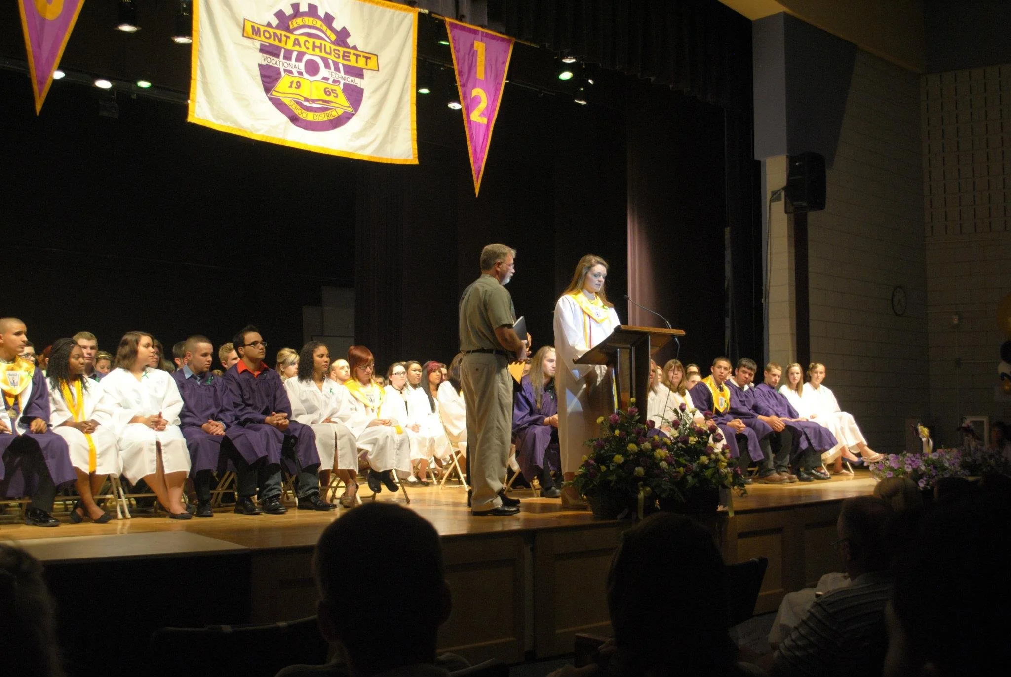 Students dressed in graduation gowns and caps seated on stage during a graduation ceremony, with a woman speaking at a podium and banners hanging above.