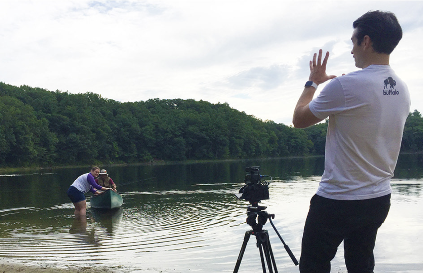 A person filming two women paddling a canoe on a river with a camera on a tripod, with lush green trees and cloudy sky in the background.