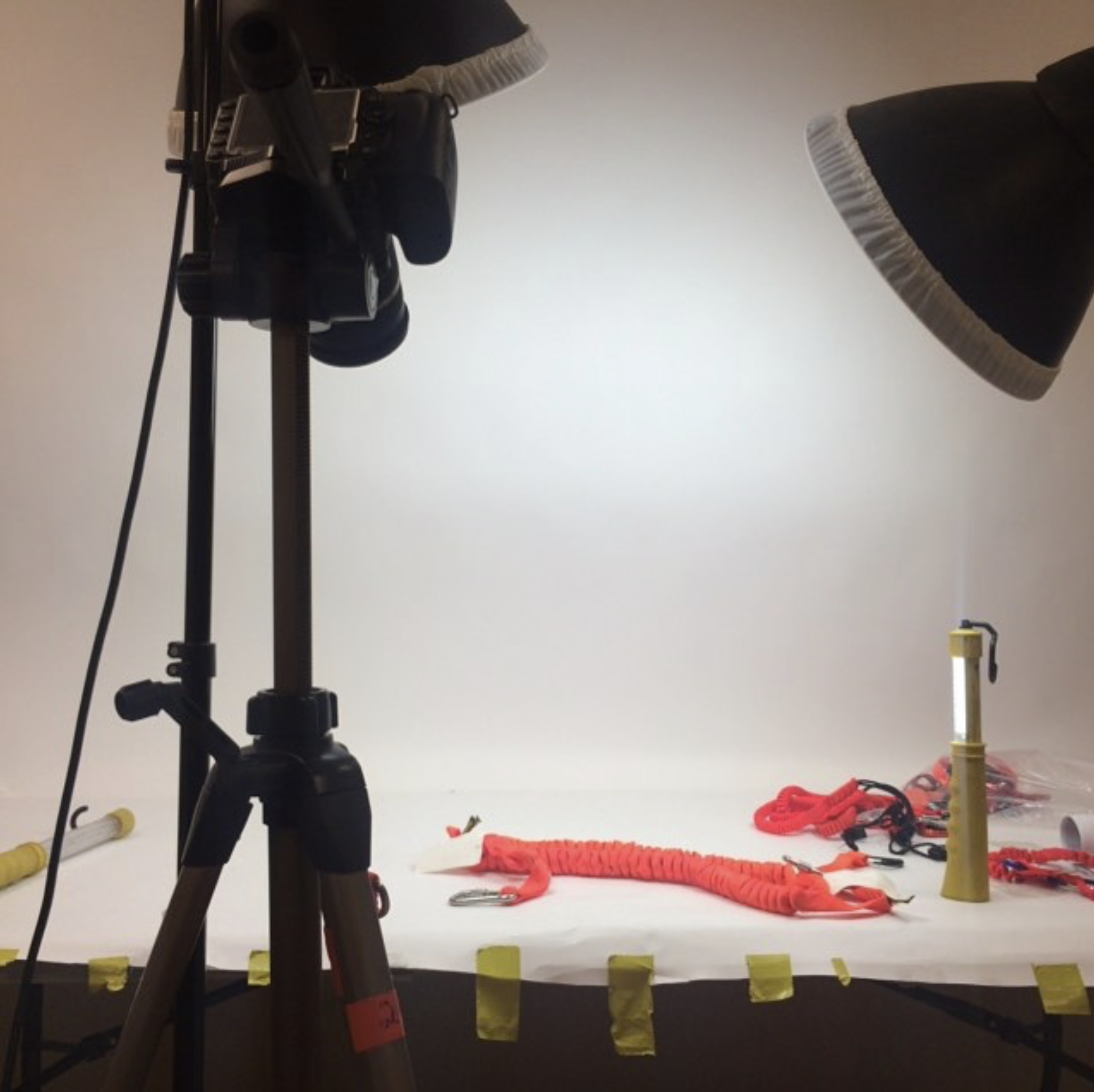 A photography setup with a black tripod, a couple of studio lights, and various equipment including red and black straps, and a yellow canister on a white background marked with yellow tape.