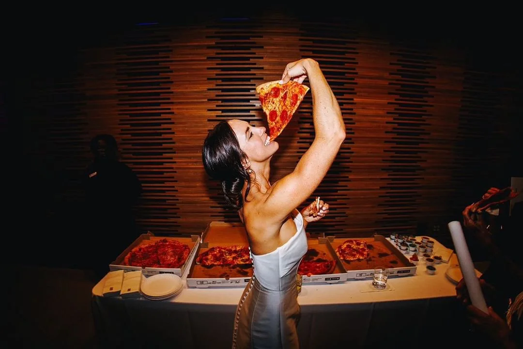Woman in white dress eating a slice of pepperoni pizza at a pizza party, with several pizza boxes on the table behind her.