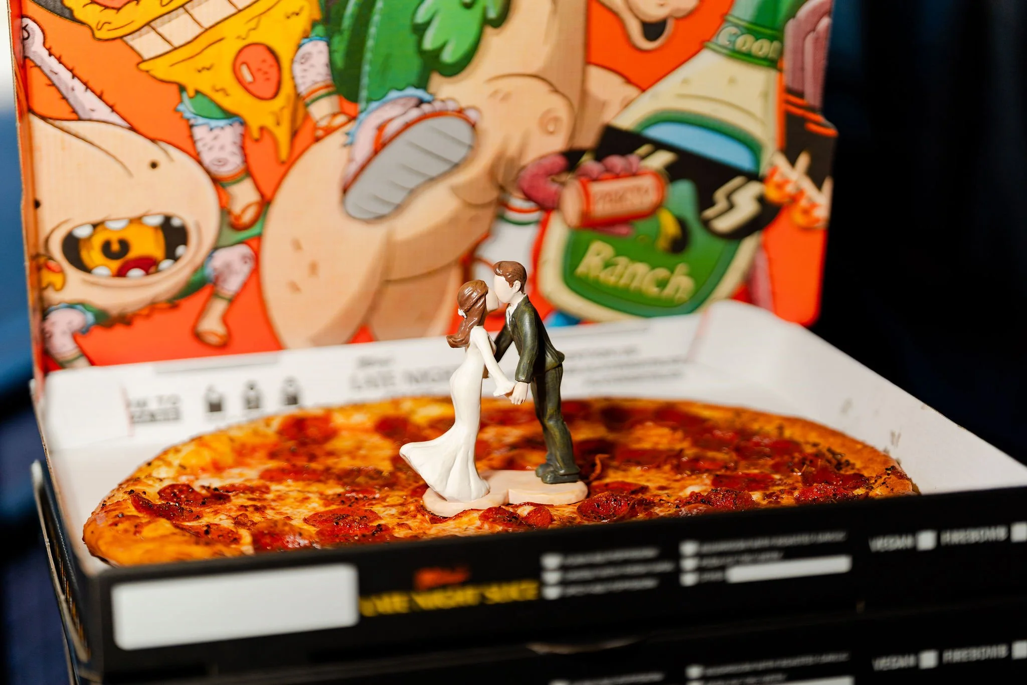 A pizza box with a pizza and plastic figurines of a man and woman dressed as a bride and groom standing on the pizza, creating a wedding scene.