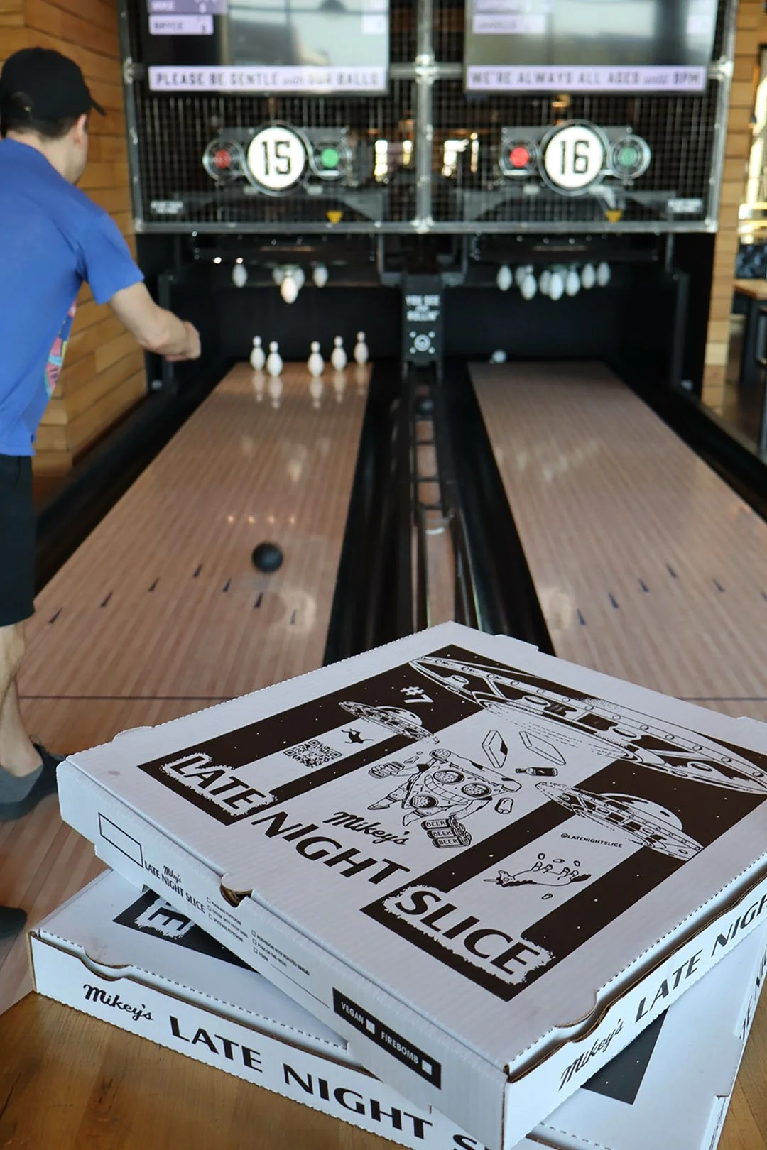 Two boxes of Mikey's Late Night Slice pizza on a table in a restaurant with a shuffleboard game in the background. One person is playing the game, and the pizza boxes are in the foreground.
