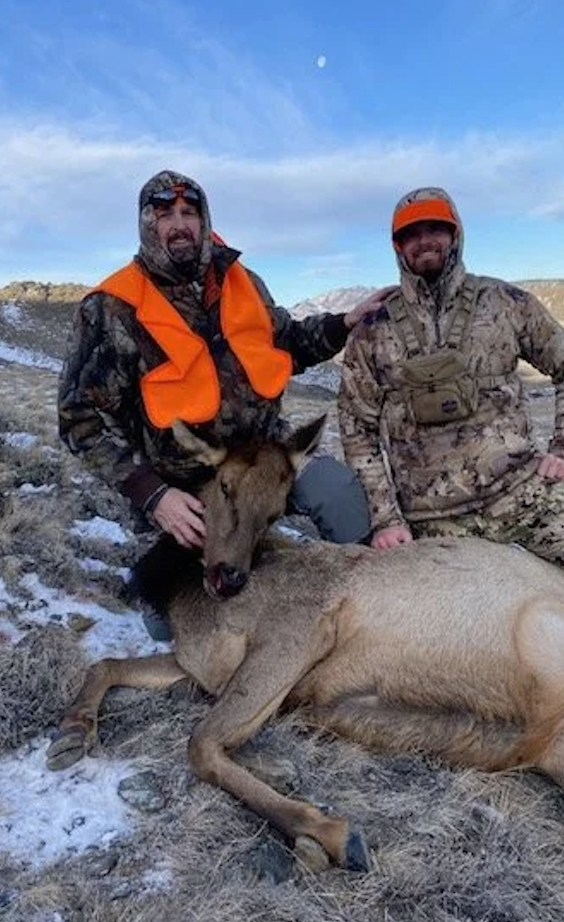 Two hunters in camouflage gear with orange vests and hats pose outdoors with a freshly hunted elk in a snowy field, one holding the animal's head.