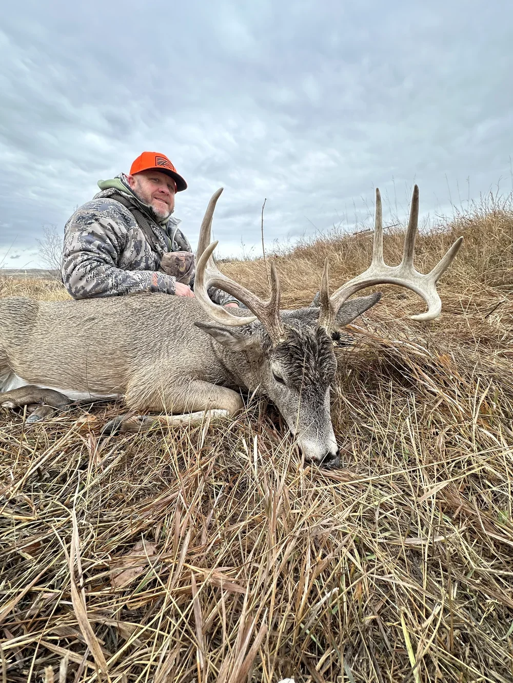 A man in camouflage clothing and orange cap kneels next to a large harvested deer with antlers, lying on dry grass under a cloudy sky.