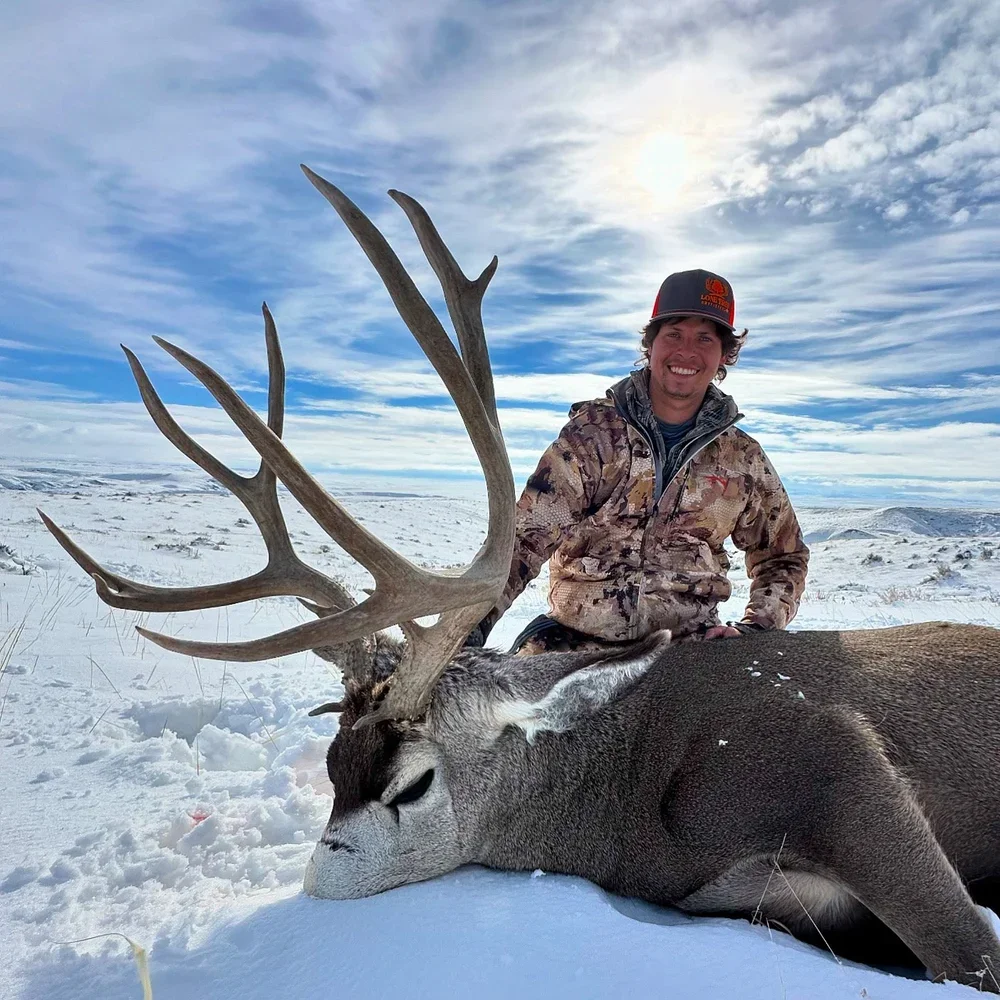 A man dressed in camouflage clothing and a cap is kneeling behind a large, freshly killed deer with impressive antlers, lying on snow in an open, snowy landscape with cloudy skies.