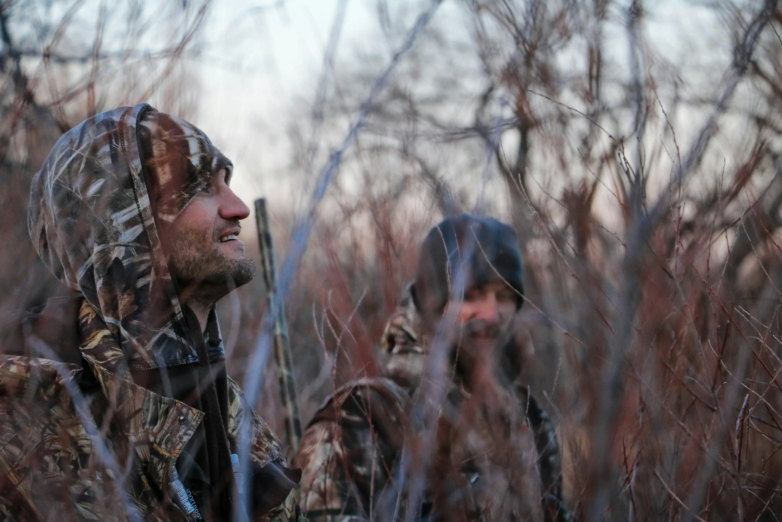 Two men wearing camouflage jackets and hoods standing among leafless bushes in a winter landscape