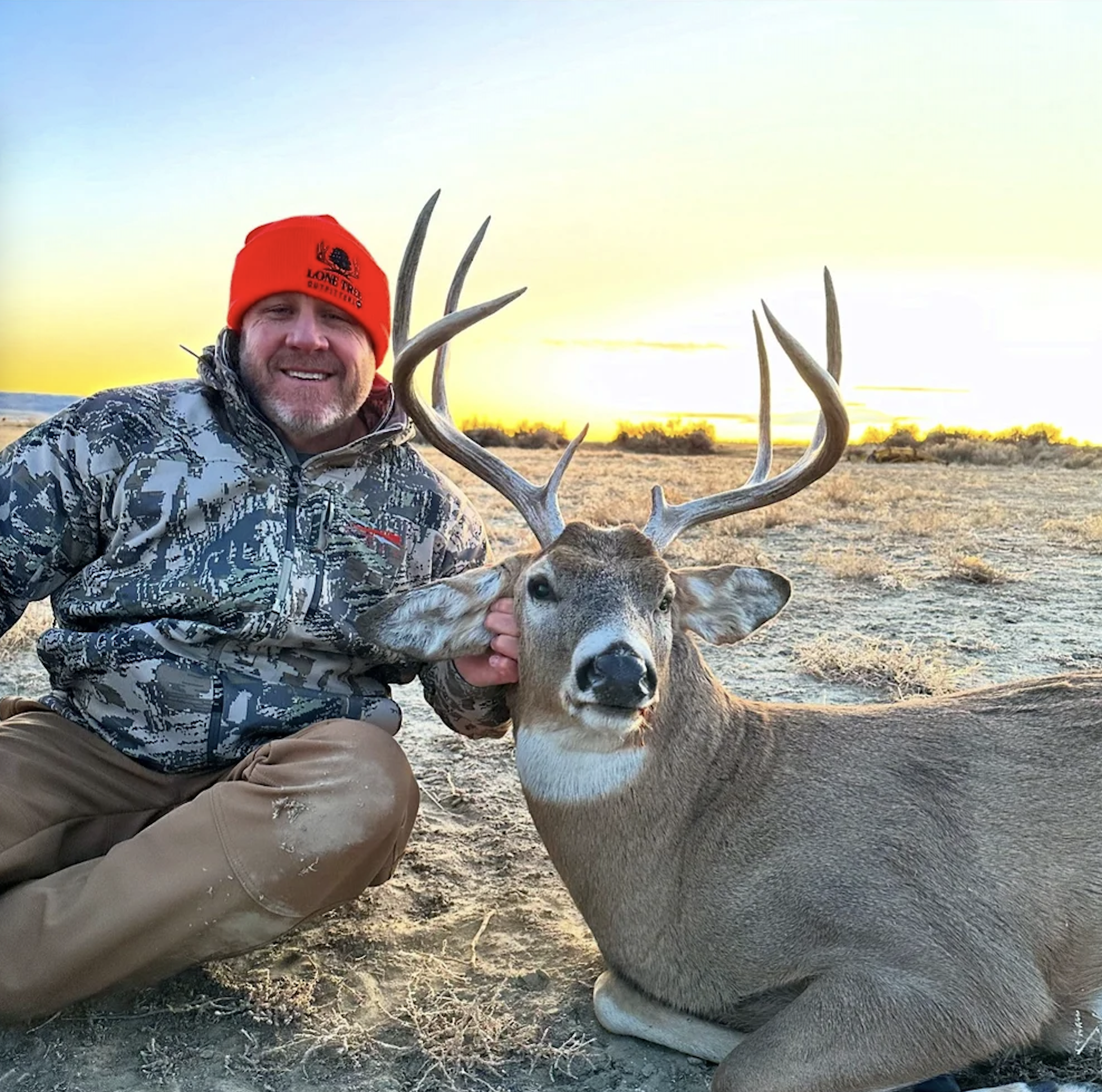 A man wearing camouflage clothing and a red hat is kneeling next to a large deer with antlers. The man is smiling and holding the deer's head, with the sunset in the background.