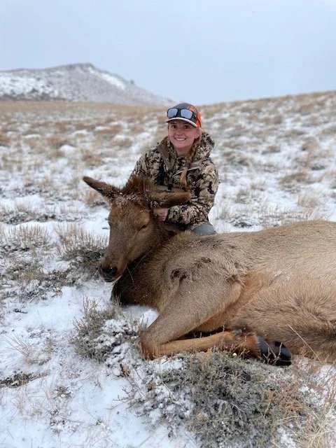 A woman in camouflage clothing and sunglasses kneels in the snow, smiling next to a dead elk lying on the ground.
