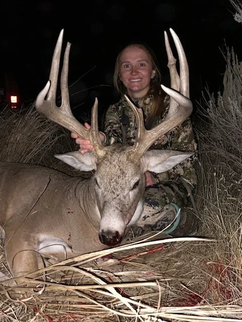 A woman in camouflage clothing holding a large deer with antlers at night, kneeling behind it in tall grass.