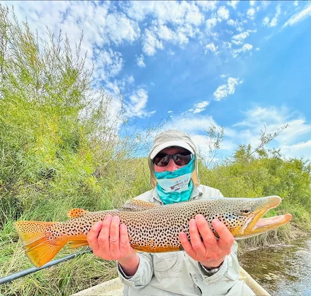 A person wearing a hat, sunglasses, and a face mask holding a large fish outdoors near water with green vegetation and a blue sky with clouds in the background.