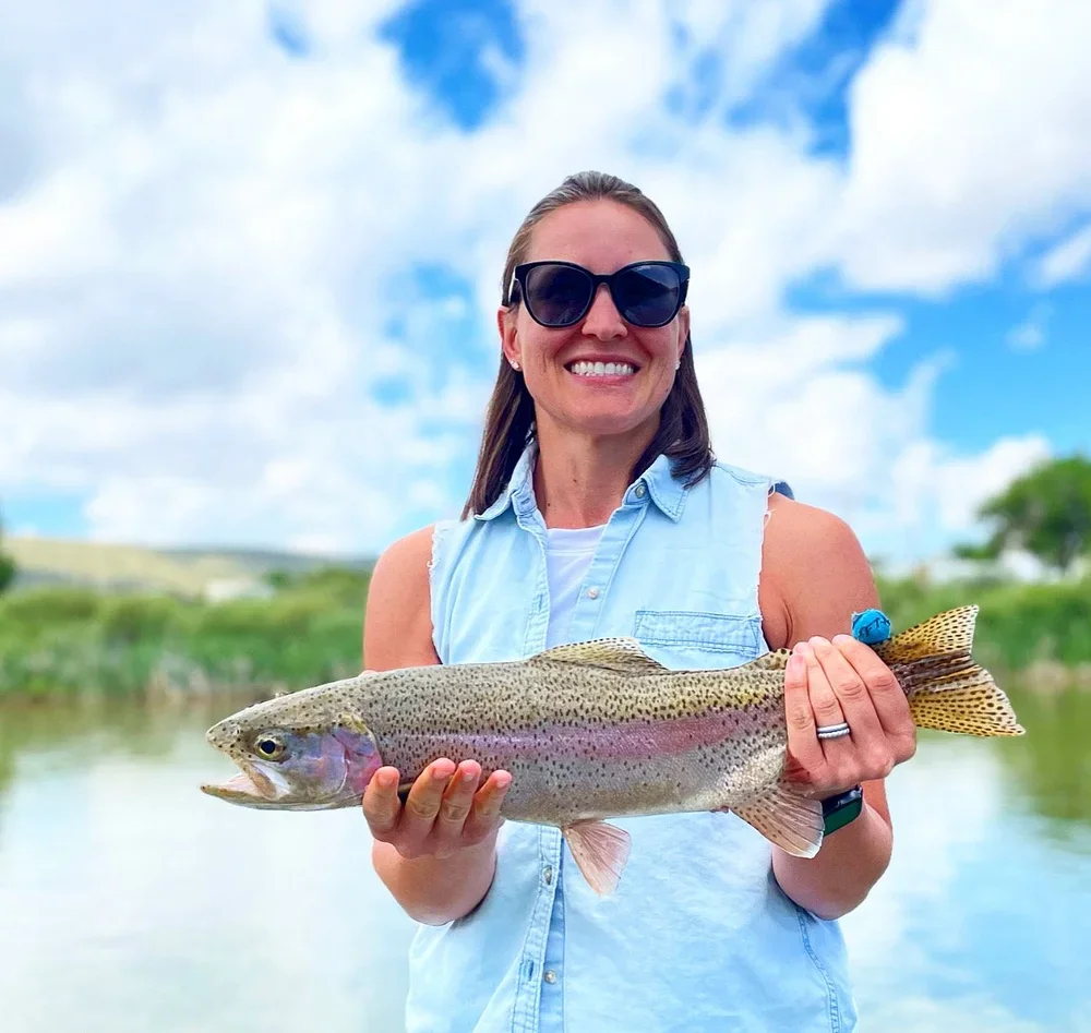 Woman in sunglasses smiling and holding a large rainbow trout outdoors with a river and cloudy sky in the background.
