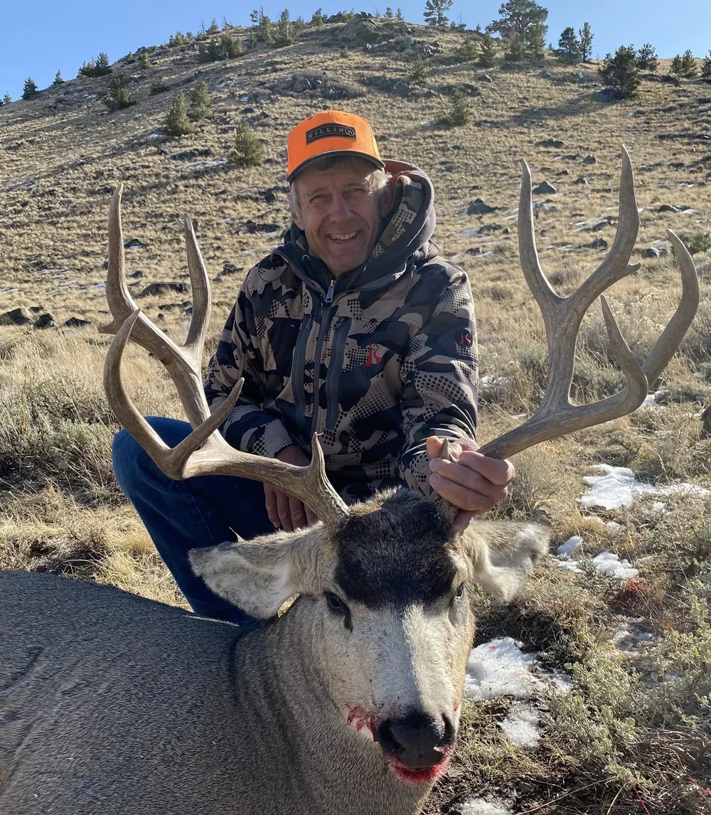 A man outdoors in a mountainous area holding the antlers of a freshly hunted elk with a laceration on its nose, wearing camouflage jacket and orange cap, smiling at the camera.