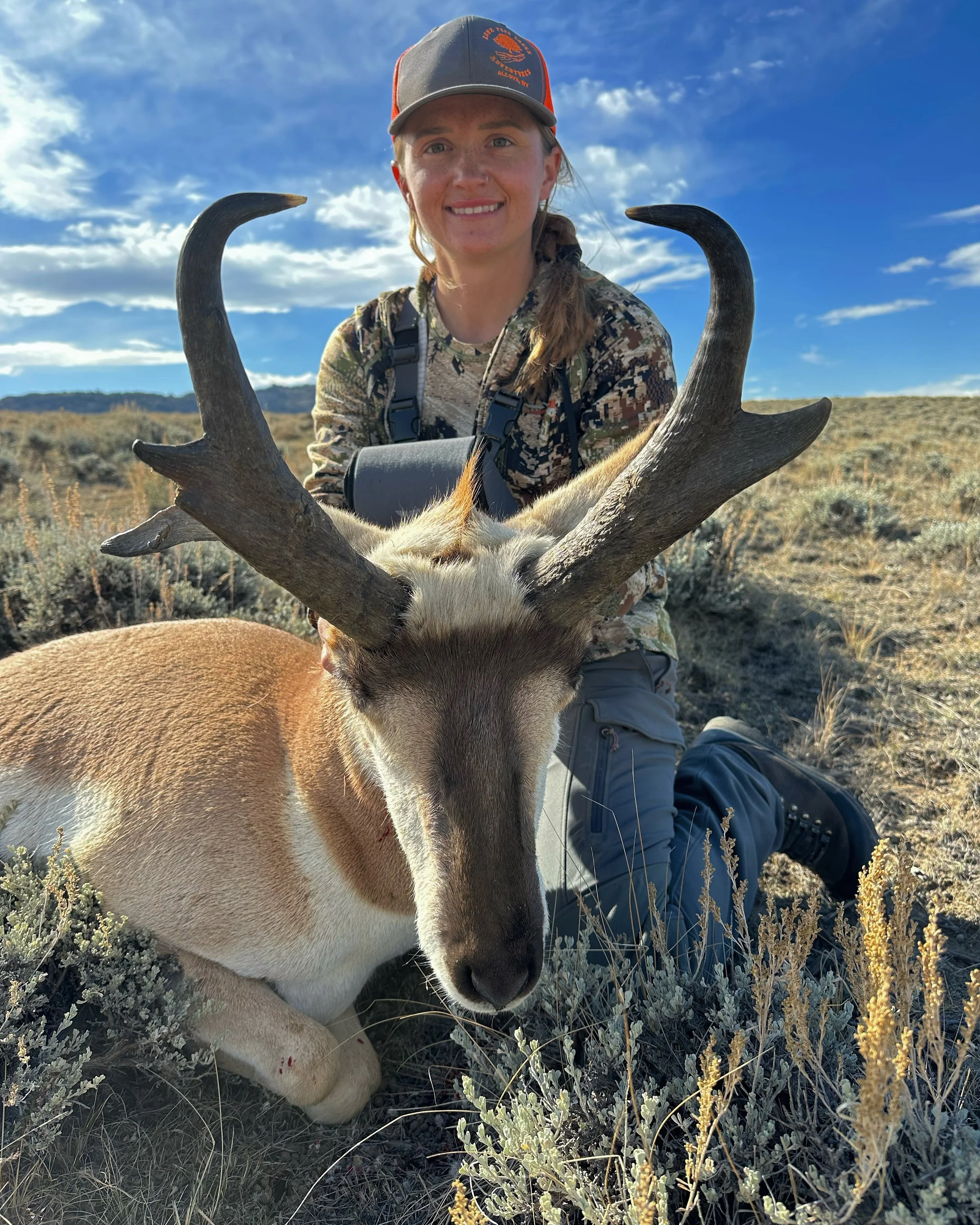 A woman kneeling on the ground in a desert-like landscape, smiling, with a large deer with antlers in front of her.