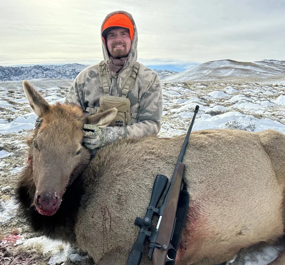 Man dressed in camouflage clothing with a bolt-action rifle resting on a dead elk in a snowy landscape with rolling hills in the background.