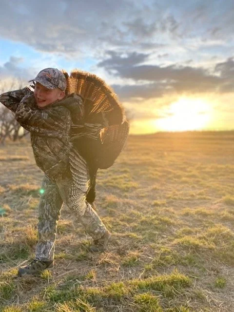 A child dressed in camouflage clothing carrying a large basket over their shoulder in an open field at sunset.