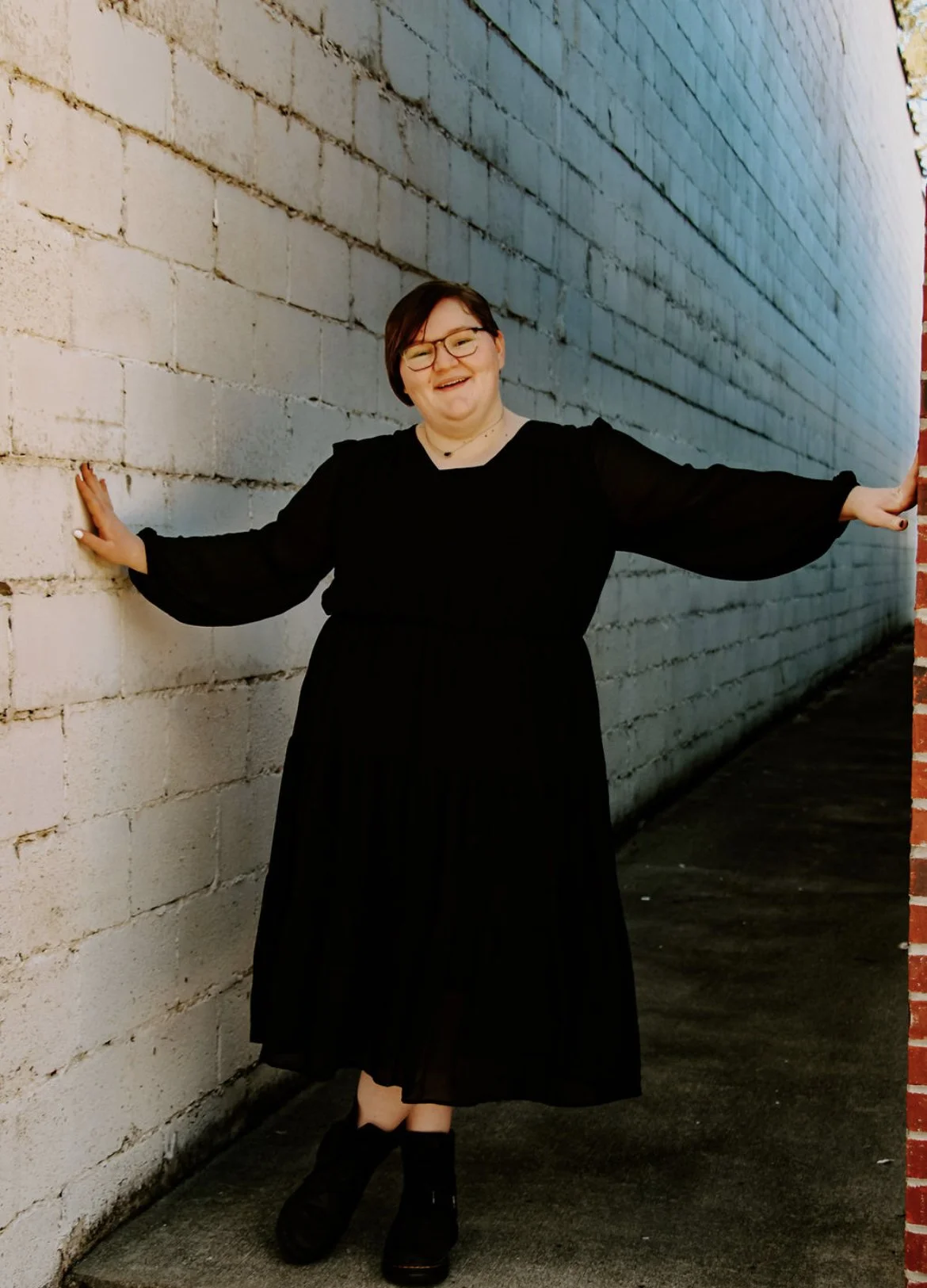Woman in black dress and glasses smiling, standing against a white brick wall with arms outstretched.