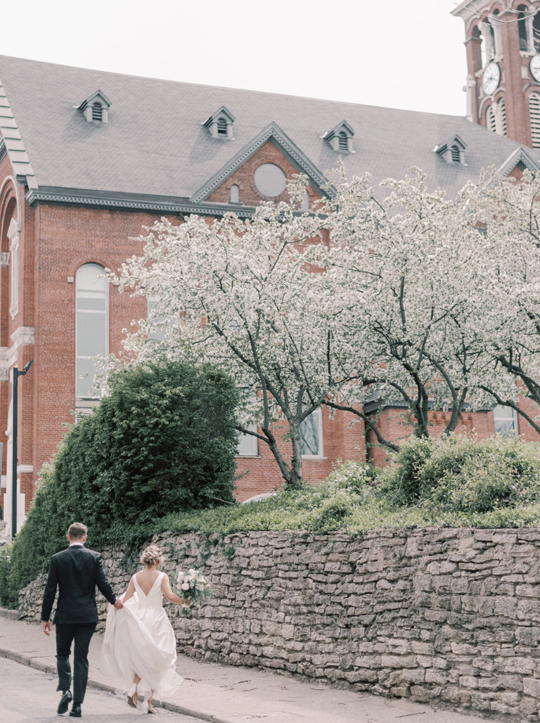A bride and groom walking hand-in-hand on a sidewalk in front of a red brick building with a clock tower, surrounded by blooming trees and green bushes.