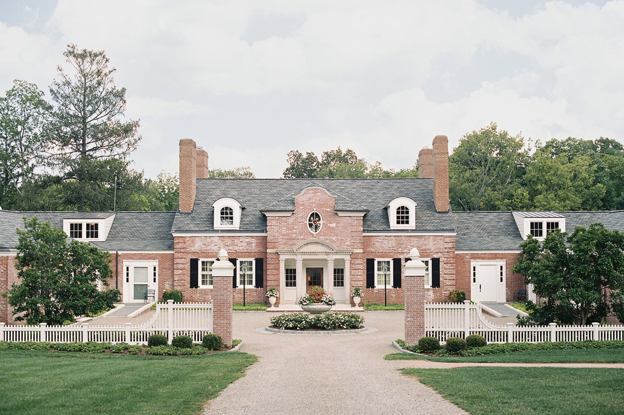 Large brick house with black shutters, a circular driveway, white picket fence, and manicured lawn and gardens.