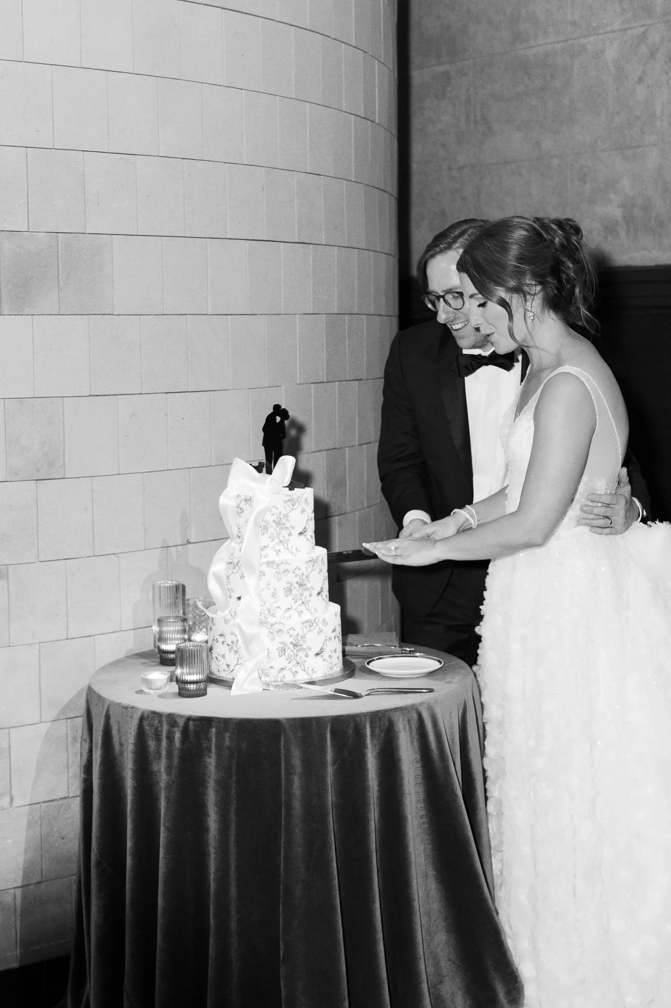 A bride and groom cutting their wedding cake together at reception, indoor setting with brick wall.