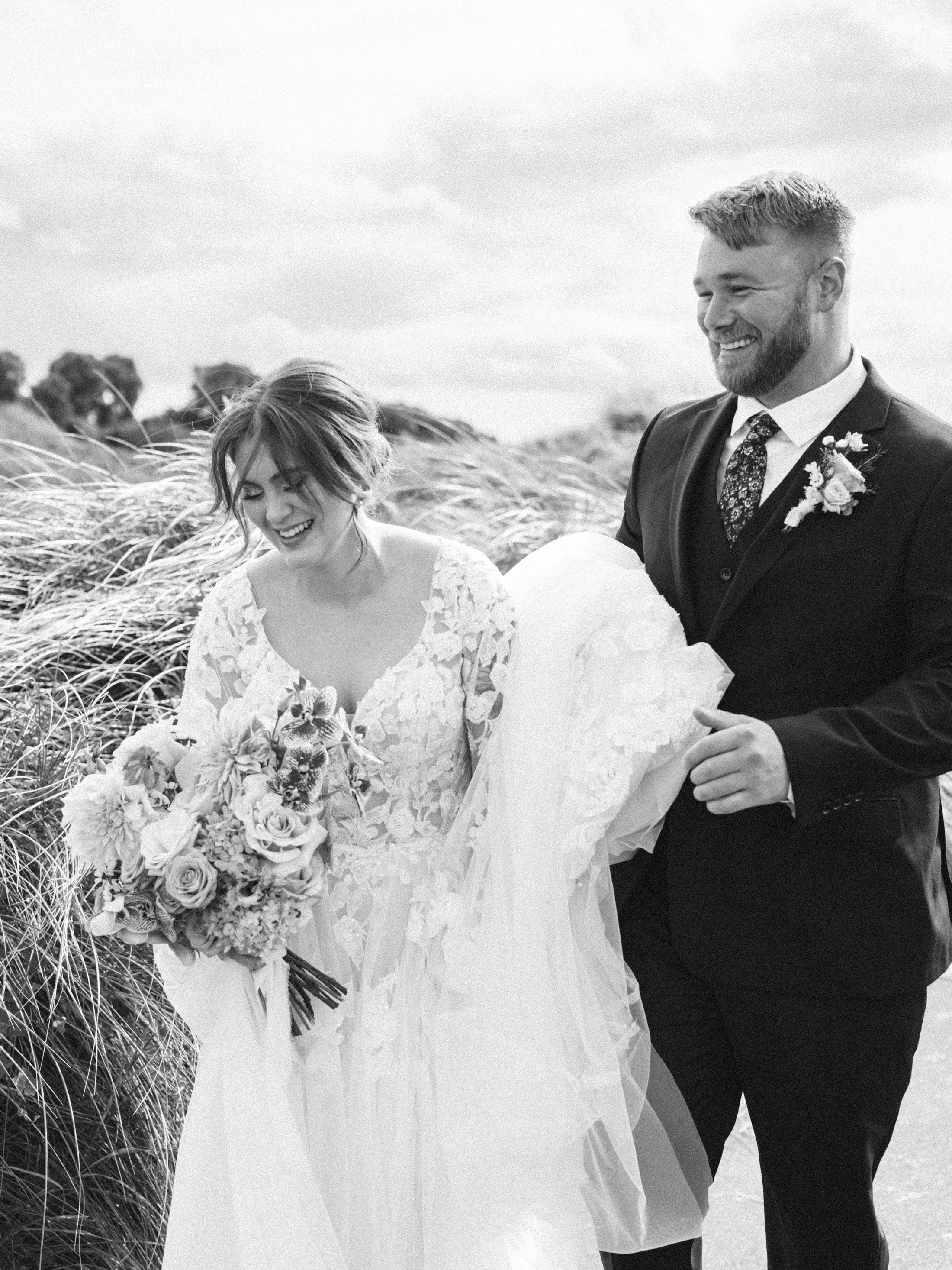 A bride in a lace wedding dress holding a bouquet of flowers and a groom in a suit smiling outdoors with tall grass in the background.