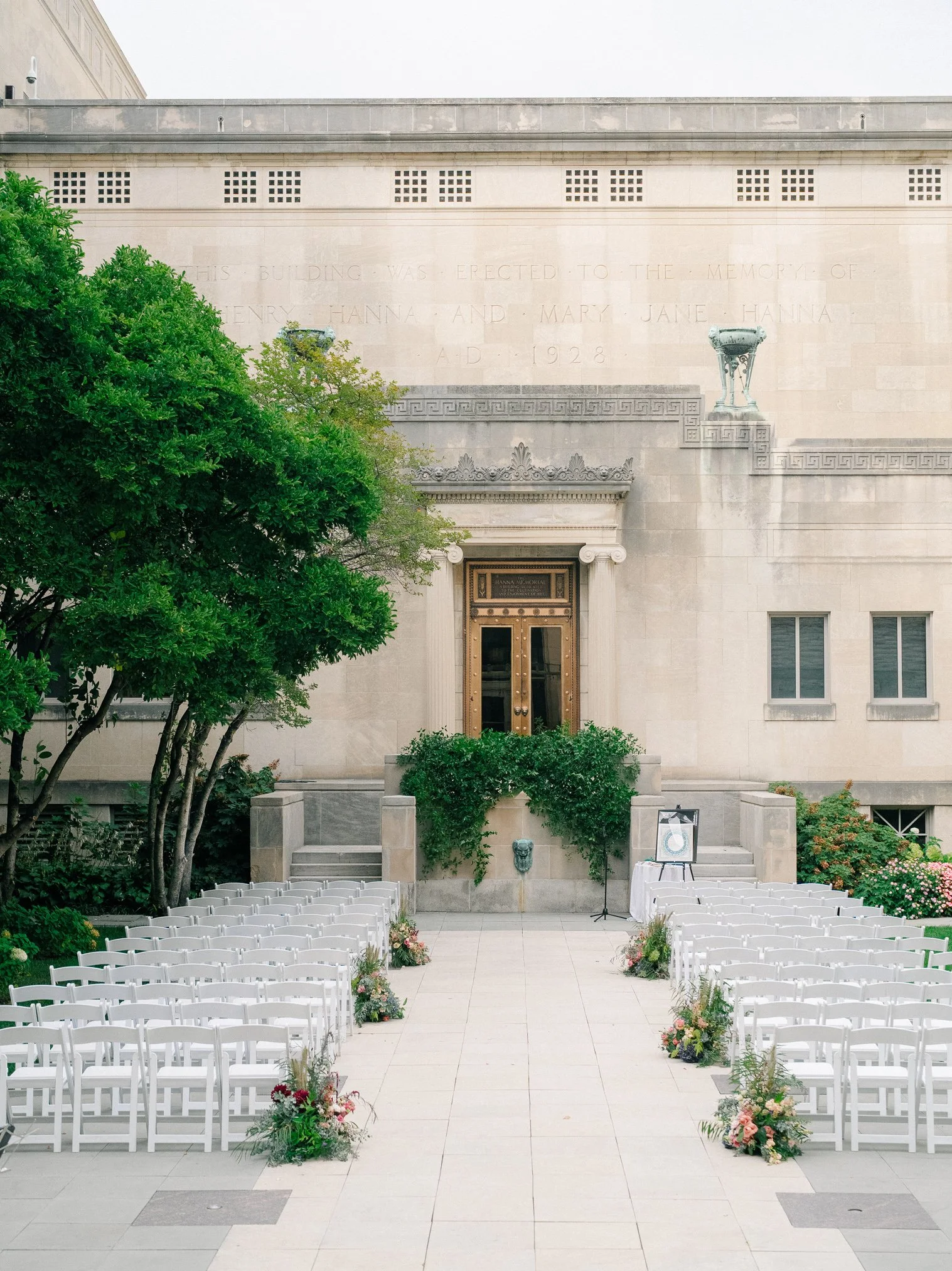 Outdoor wedding setup with white chairs and floral arrangements facing a building with steps and a door, greenery to the sides, and a stone wall with inscriptions.