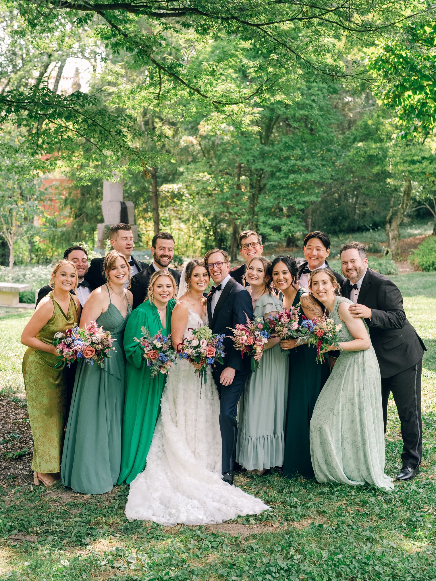 A wedding party standing outside in a park, smiling and holding bouquets.