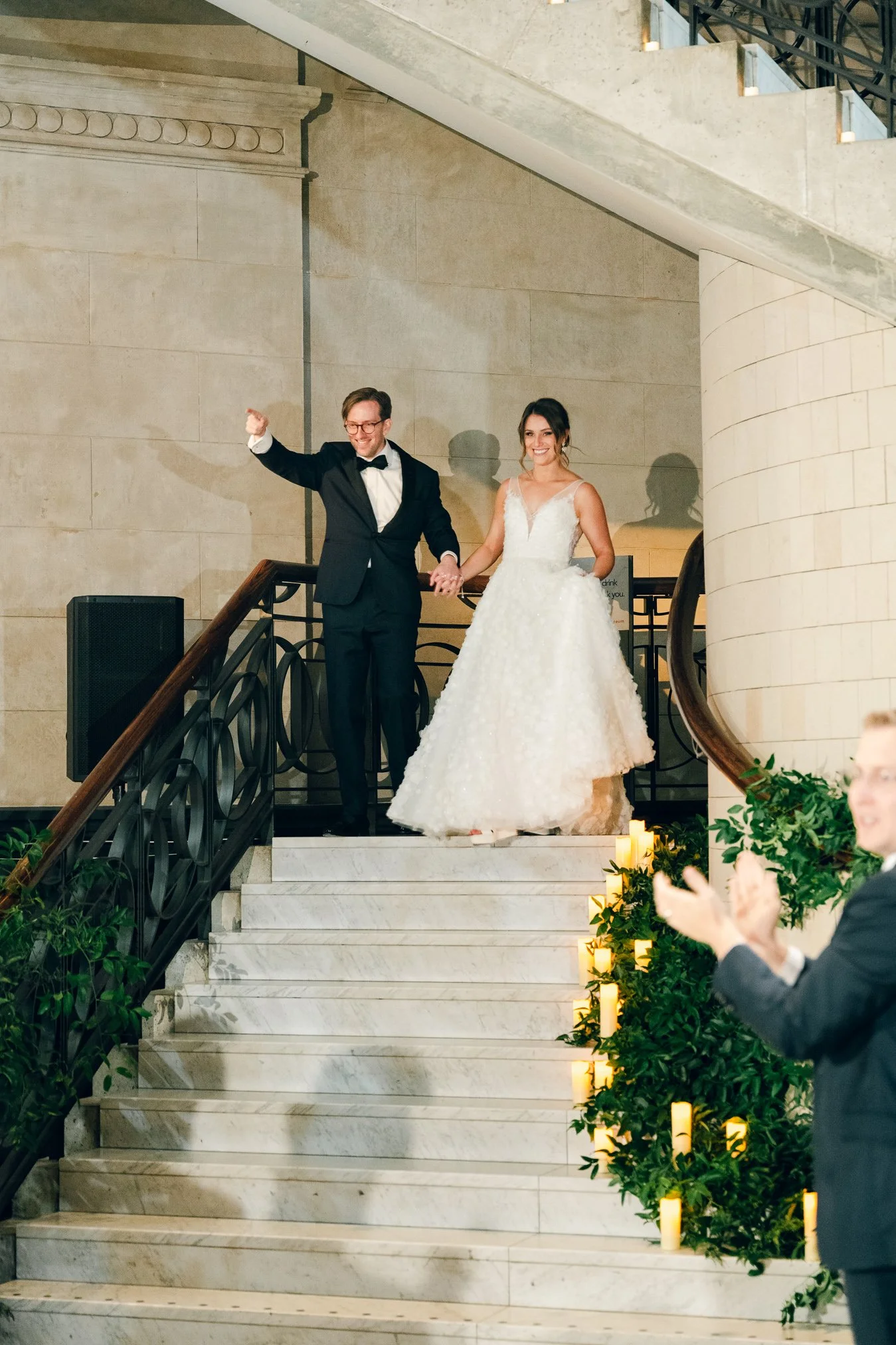 Newlyweds holding hands and celebrating at their wedding reception, standing on a staircase inside a large venue with candles and greenery decoration.