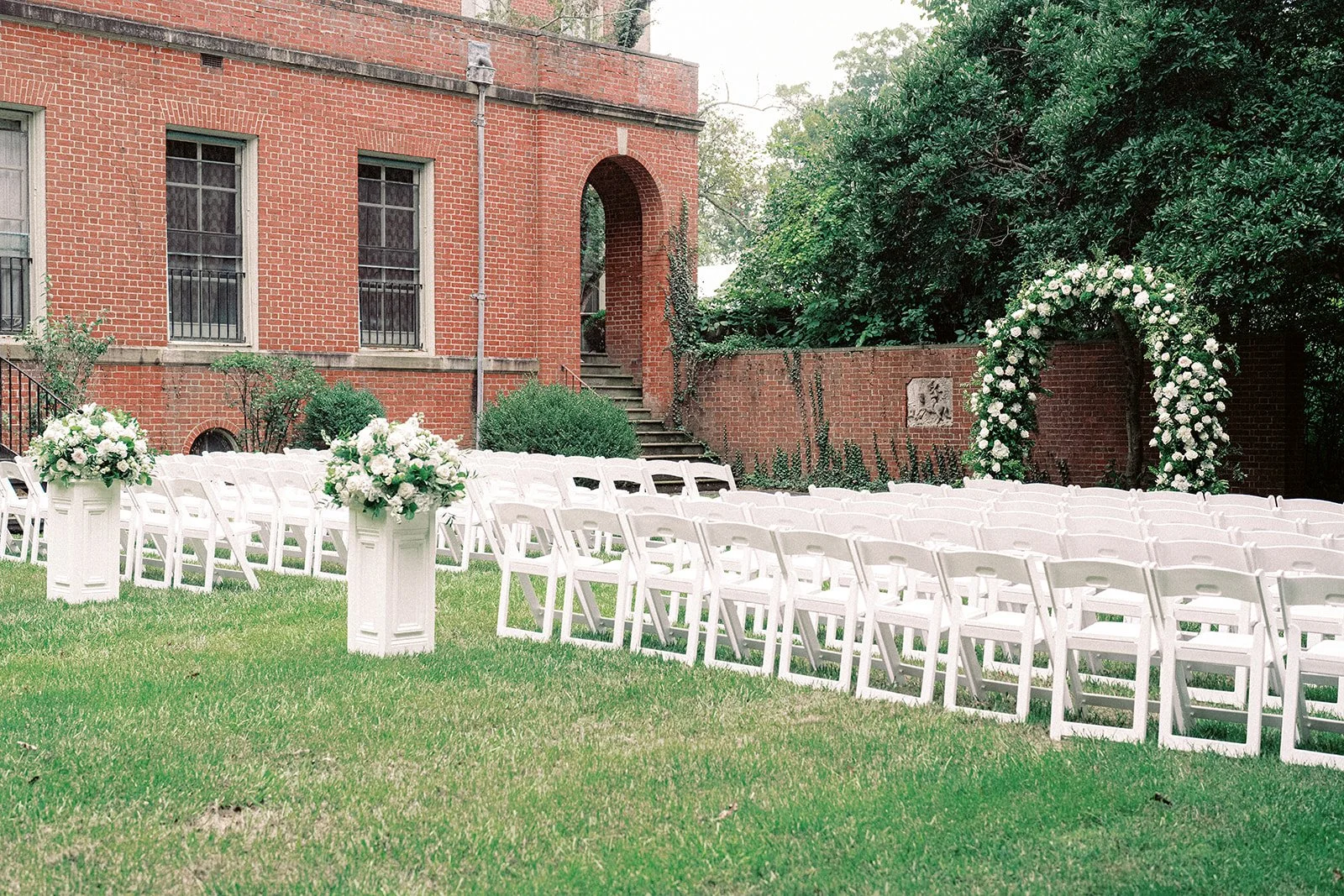 Wedding Garden ceremony outdoors at Peterloon Estate in Cincinnati. White garden chairs, floral arrangements, and an arch, in front of a brick wall and building.