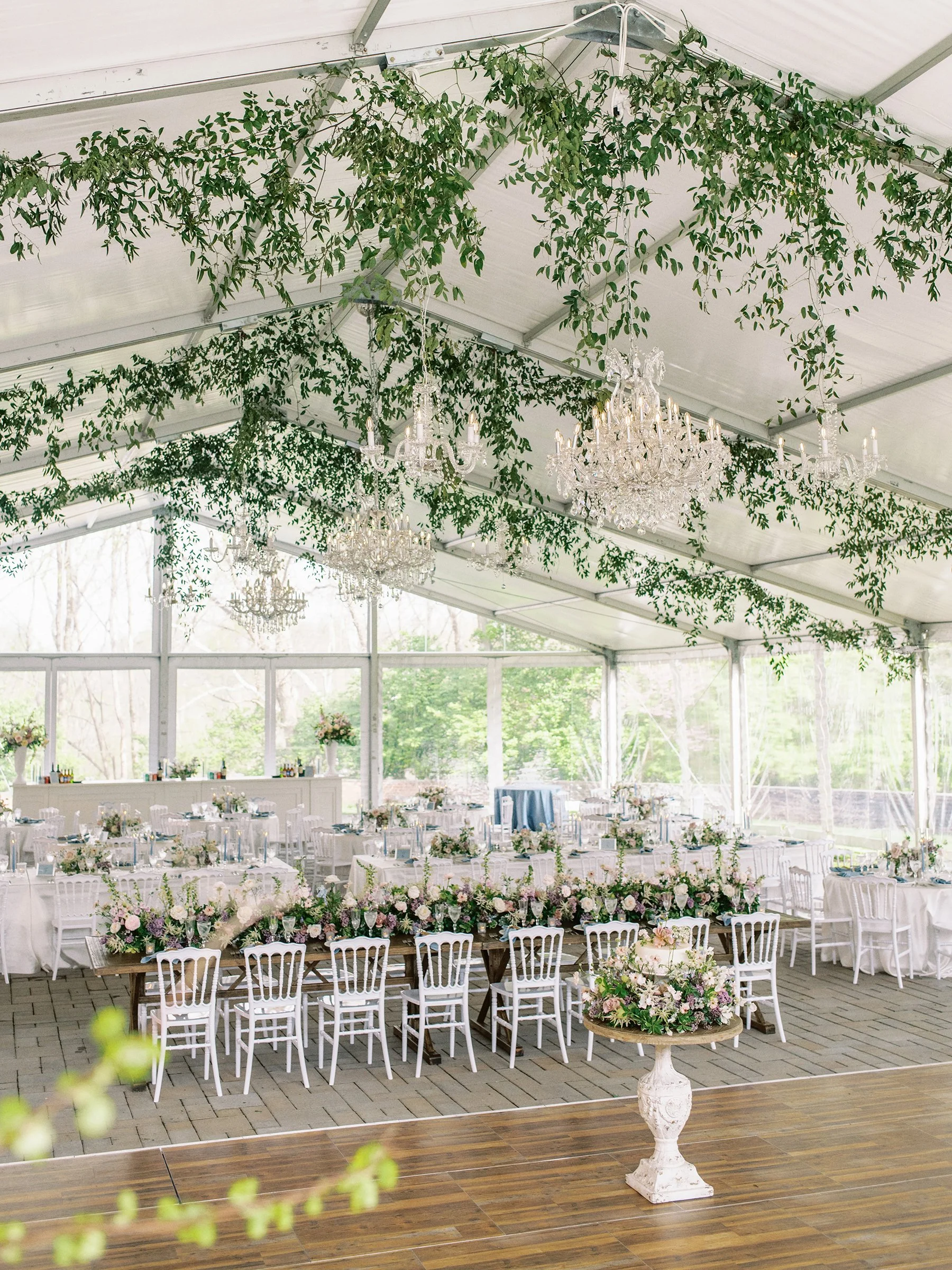 Elegant wedding reception setup with white chairs and tables, floral centerpieces, crystal chandeliers, and greenery hanging from the ceiling inside a tent.