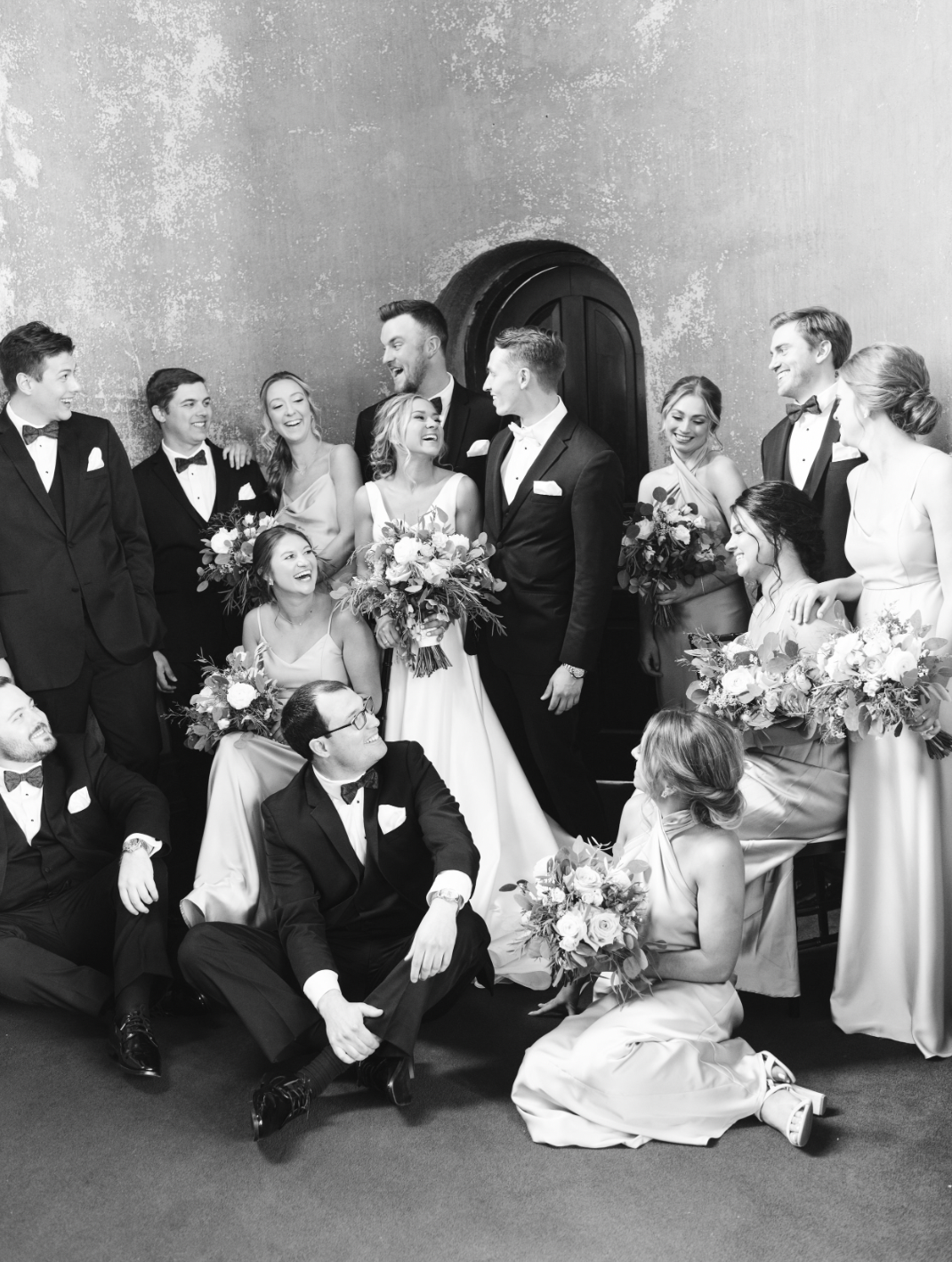 A black and white photo of a wedding party with flower bouquets, including the bride and groom, surrounded by friends and family in formal attire, smiling and laughing together.