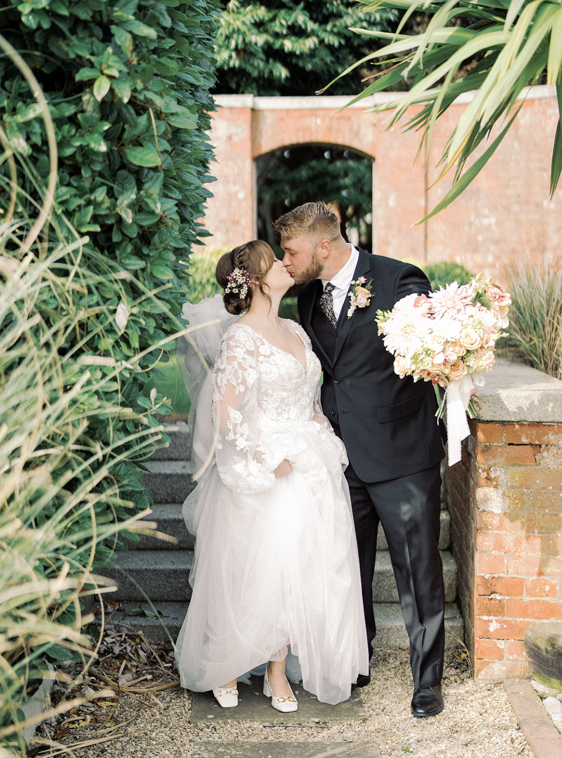 Bride and groom sharing a kiss outdoors, bride in white lace dress and veil, groom in black suit holding a pink and white flower bouquet, surrounded by greenery and red brick architecture.