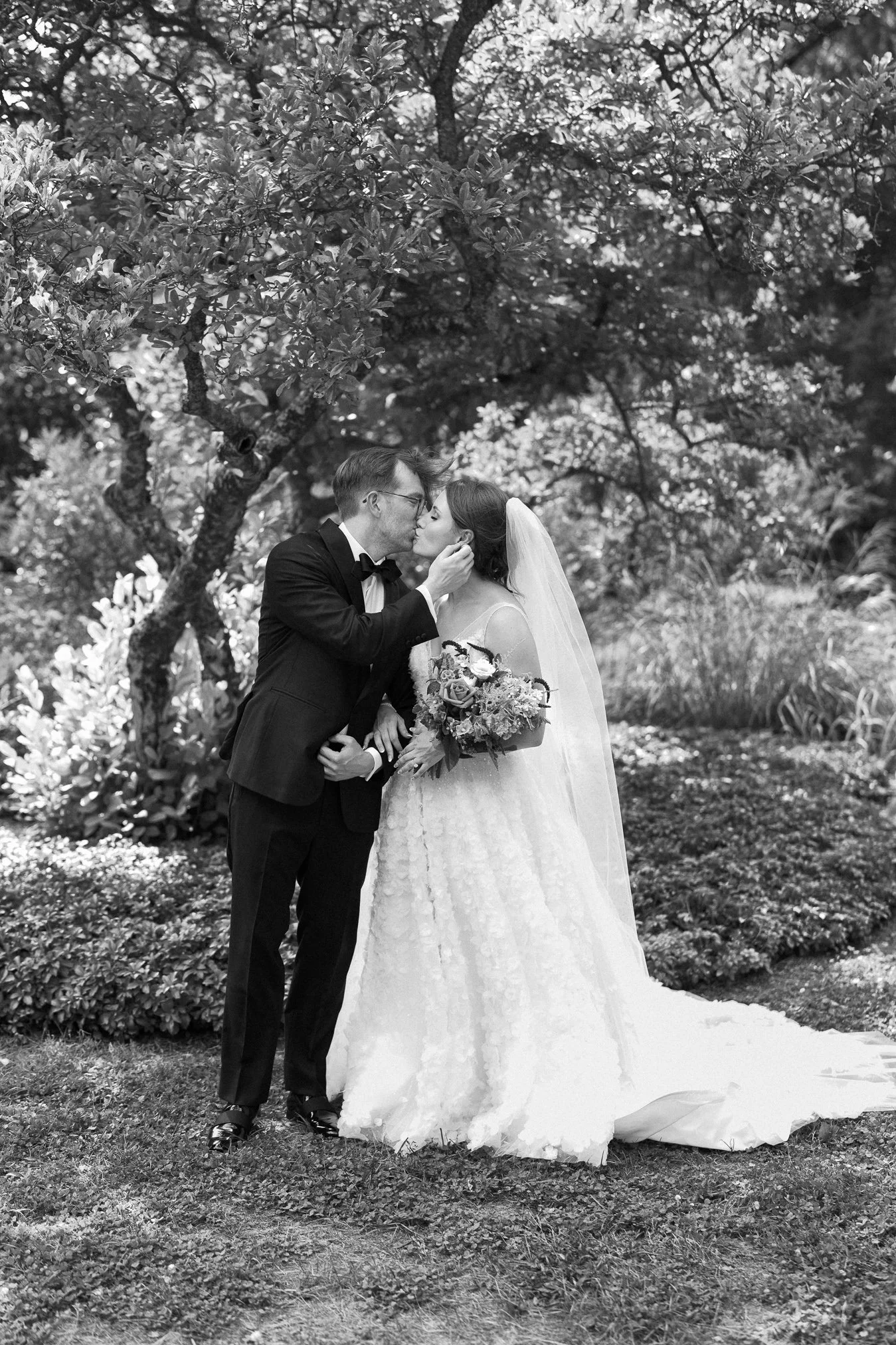 A black and white photo of a wedding couple outdoors, with the groom gently holding the bride's face and about to kiss her, surrounded by trees and foliage.