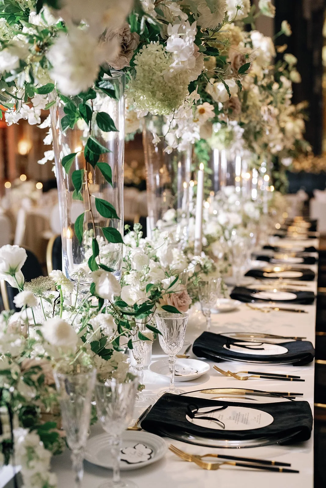 A long banquet table decorated with tall glass vases filled with white flowers and green foliage, set with black napkins, gold utensils, white plates, and elegant glassware, in a lavish, softly lit event space.