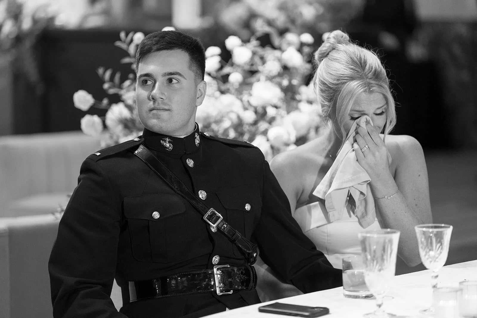 A black and white photo of a man in a military uniform sitting at a table, next to a woman wiping tears from her face with a cloth. There are empty glasses and a cellphone on the table, with a flower arrangement in the background.
