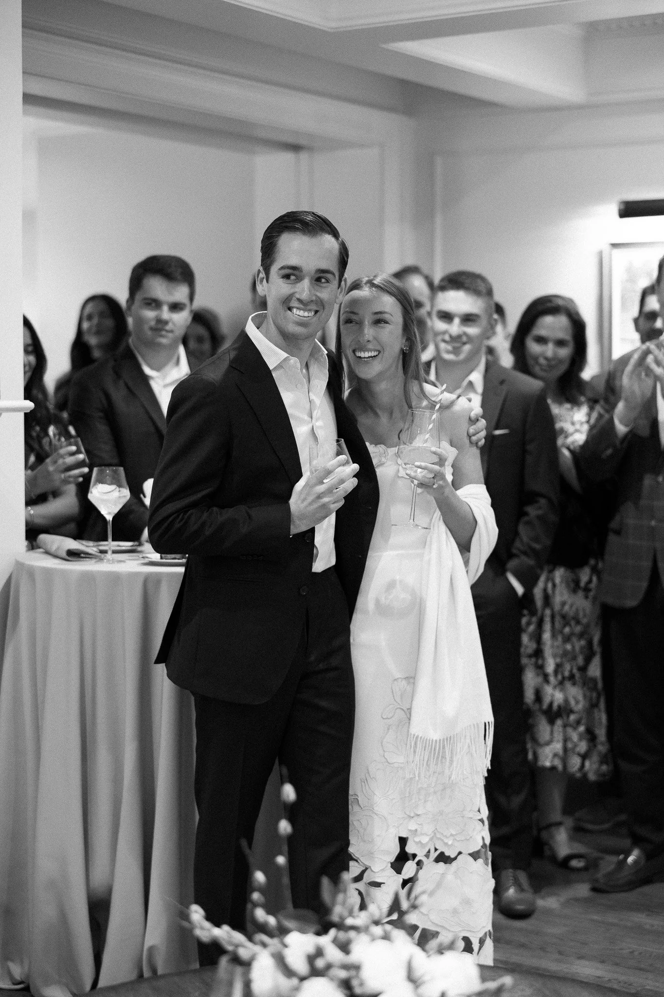 A black and white photo of a group of people at a celebration, with a man and woman in the foreground holding glasses, smiling amid an elegant indoor gathering.