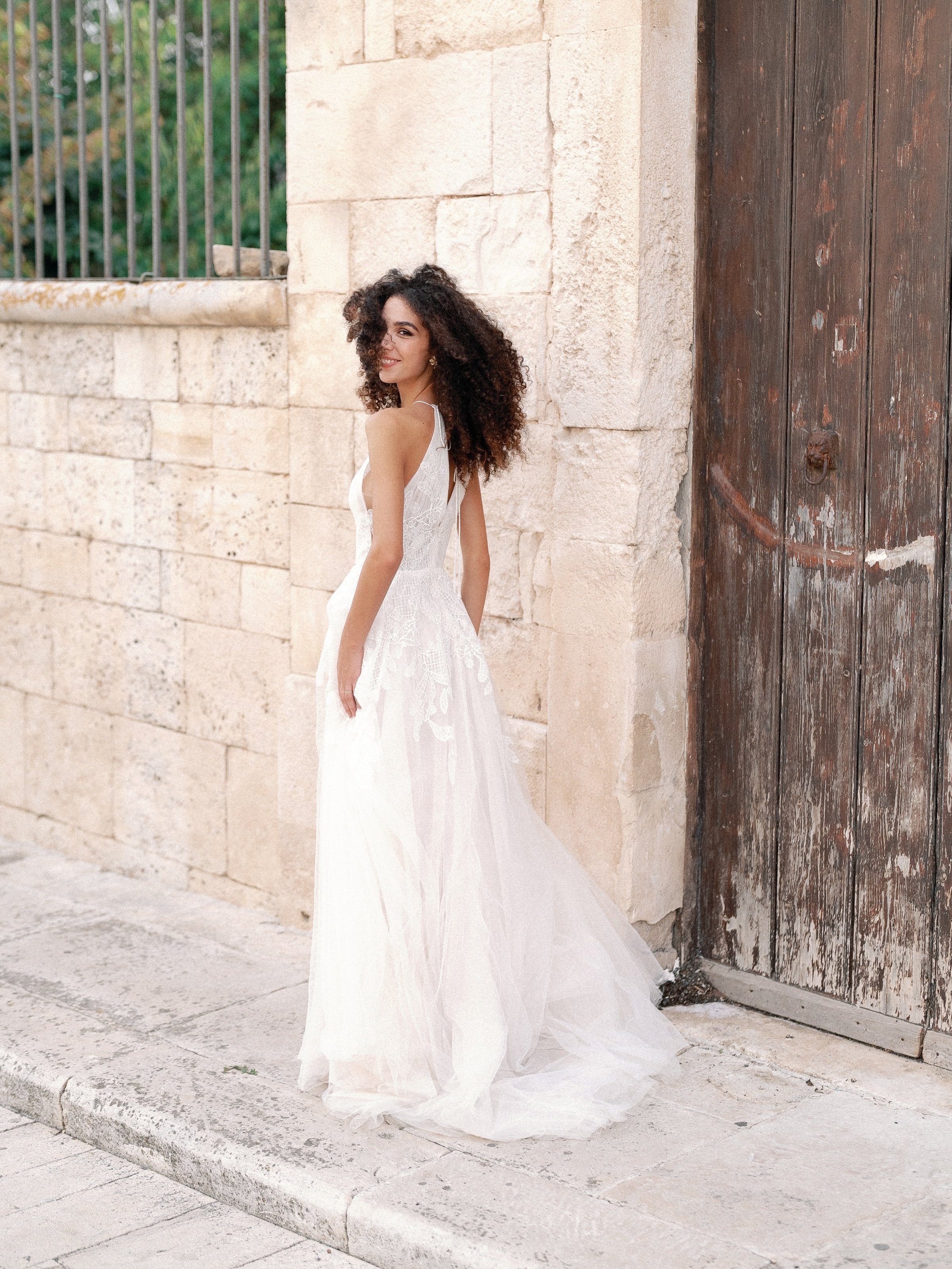 Woman in a white wedding dress standing near a stone wall and wooden door.