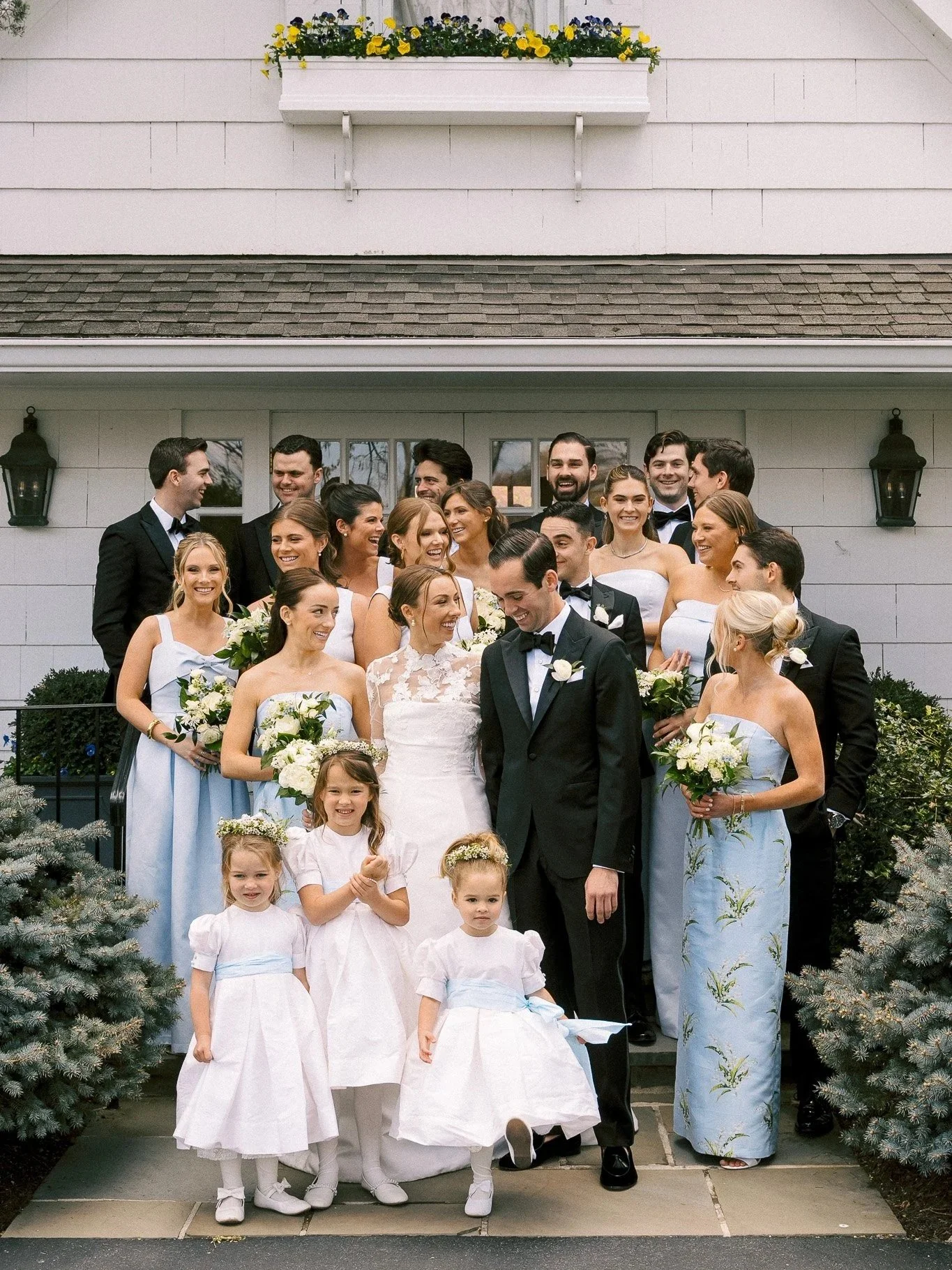 A group of people, including a bride and groom, posing for a wedding photo in front of a white house with shrubs and flowers. The bride wears a lace wedding dress, and the groom is in a black tuxedo. Several women in light blue dresses hold bouquets,
