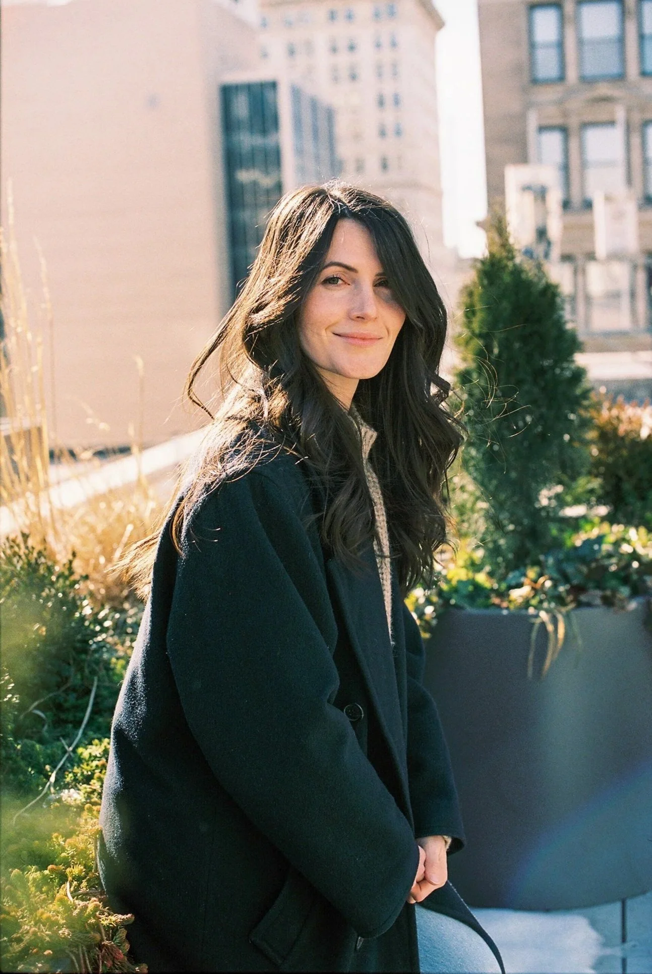 A young woman with long black hair and fair skin stands outdoors in a city, smiling slightly at the camera, with buildings and greenery in the background during daylight.