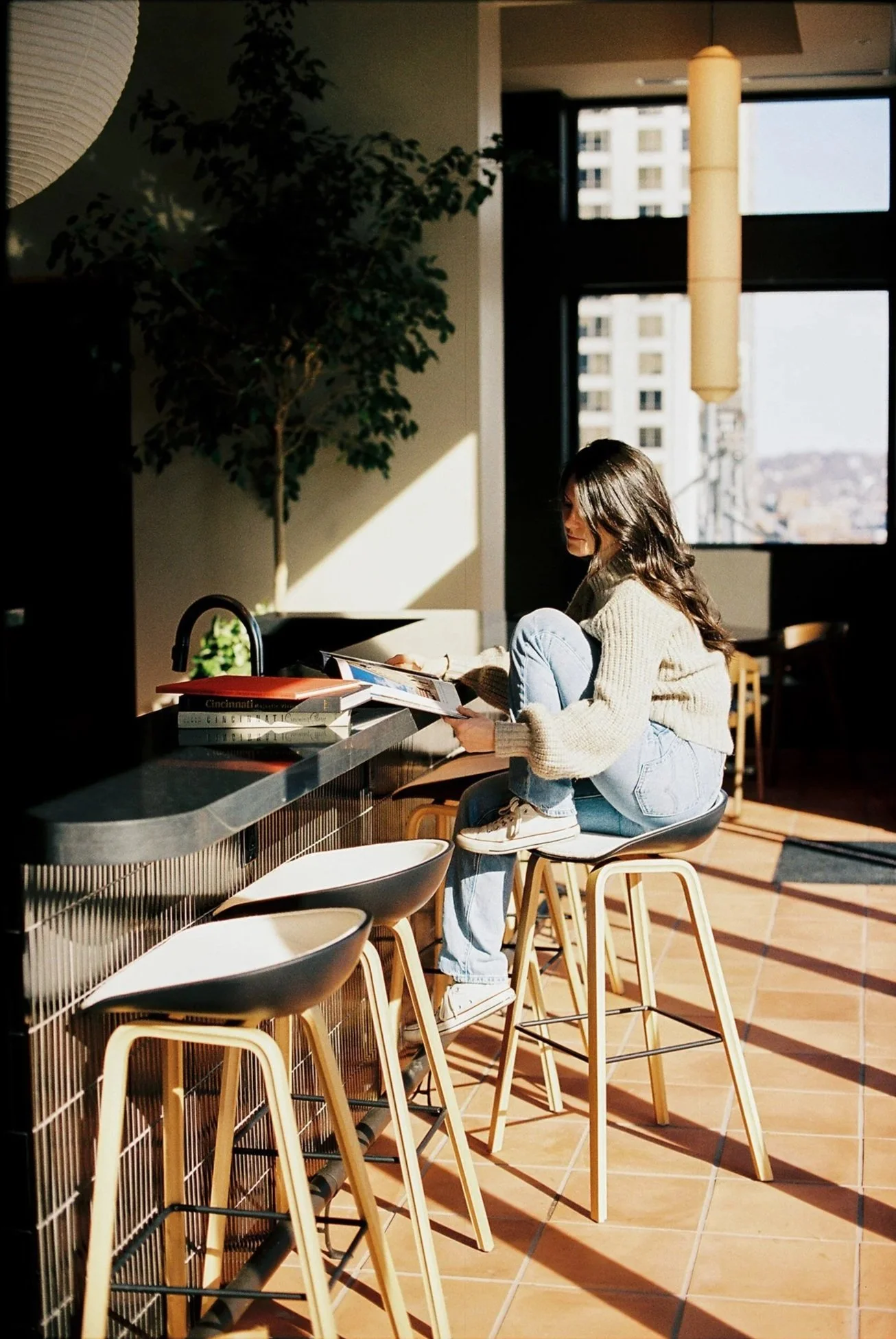 A woman sitting on a high stool at a bar counter, looking at a magazine or book, with sunlight streaming in through large windows behind her in a modern indoor space.