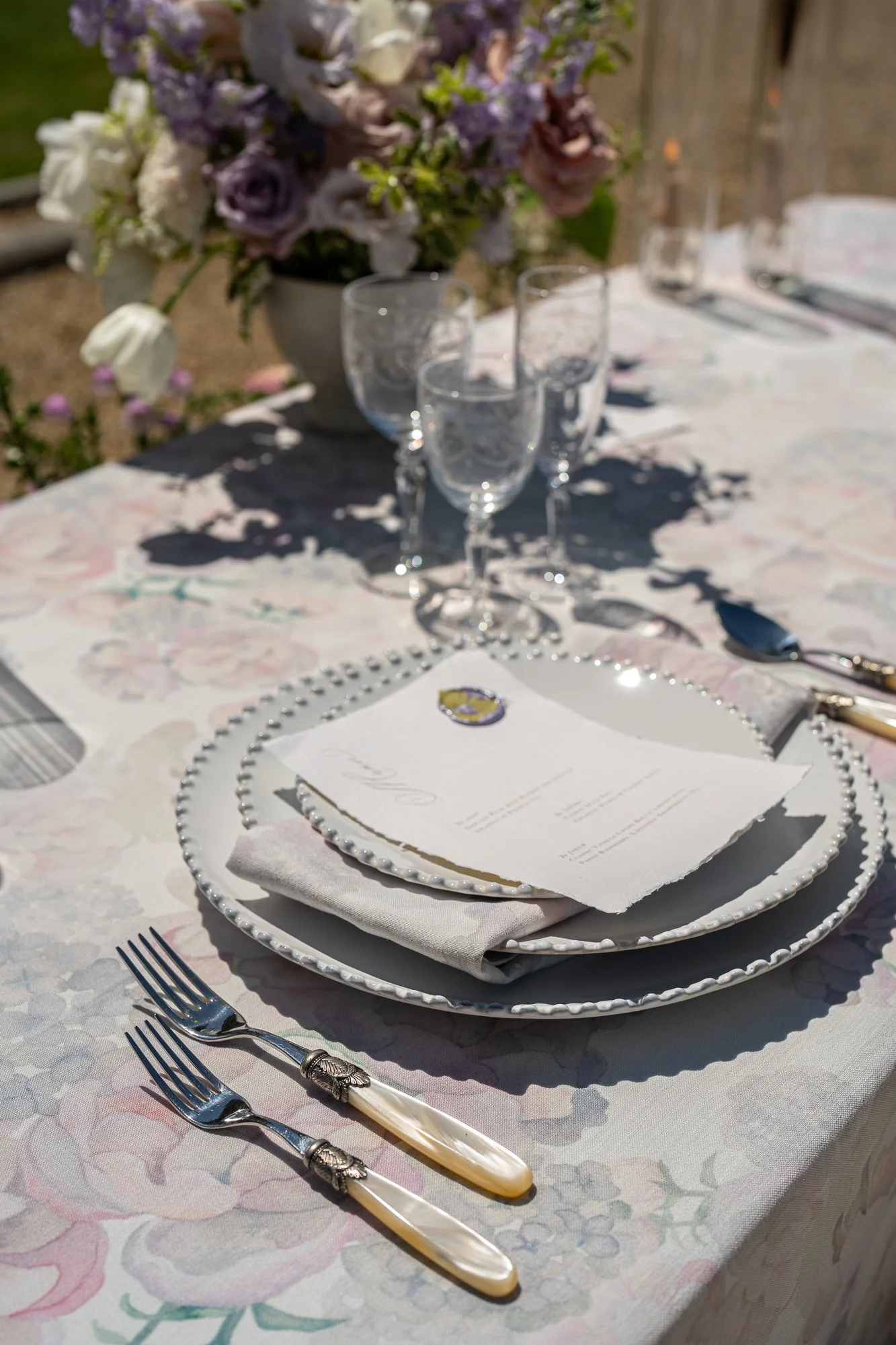A beautifully set outdoor table with floral centerpiece, wine glasses, silverware, and a menu on a decorated plate, for a formal event or celebration.