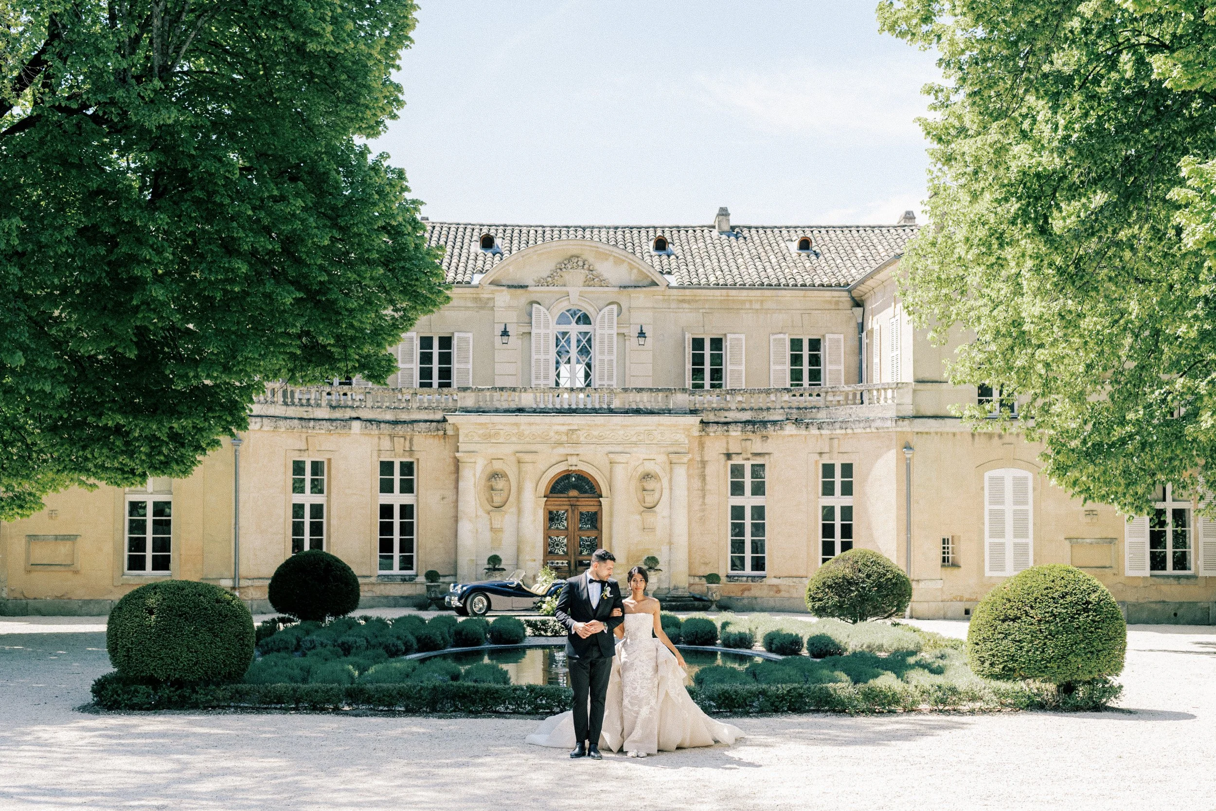 A bride and groom walking in front of a large, historic mansion with trees, bushes, and a fountain in the foreground on a sunny day.