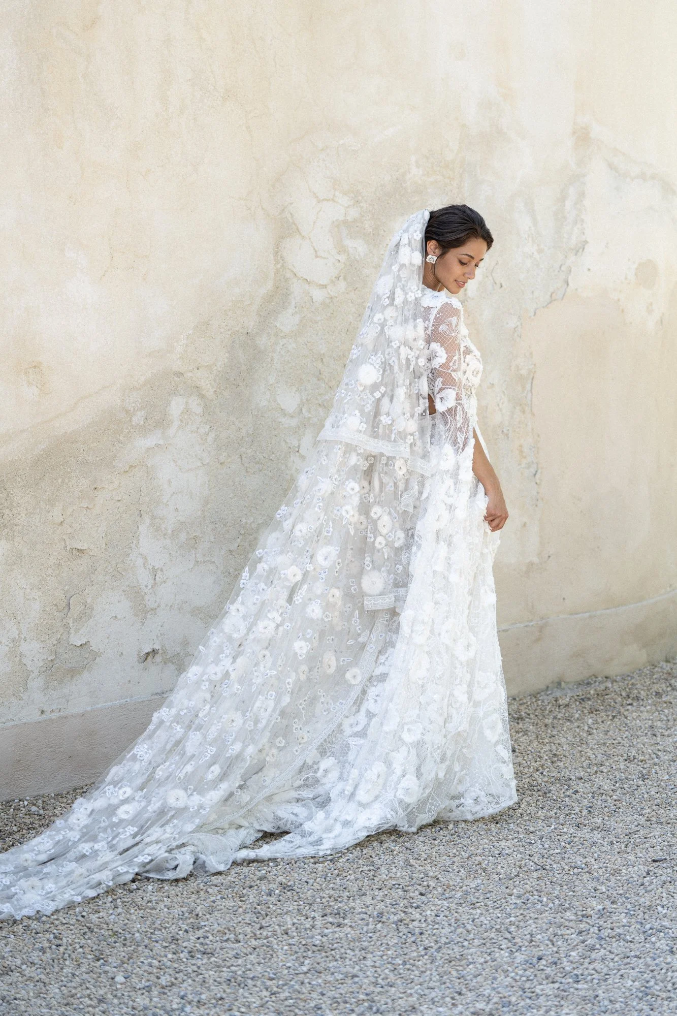 A bride in a white lace wedding dress with floral patterns and a veil, standing against a weathered beige wall with her eyes closed and smiling slightly.