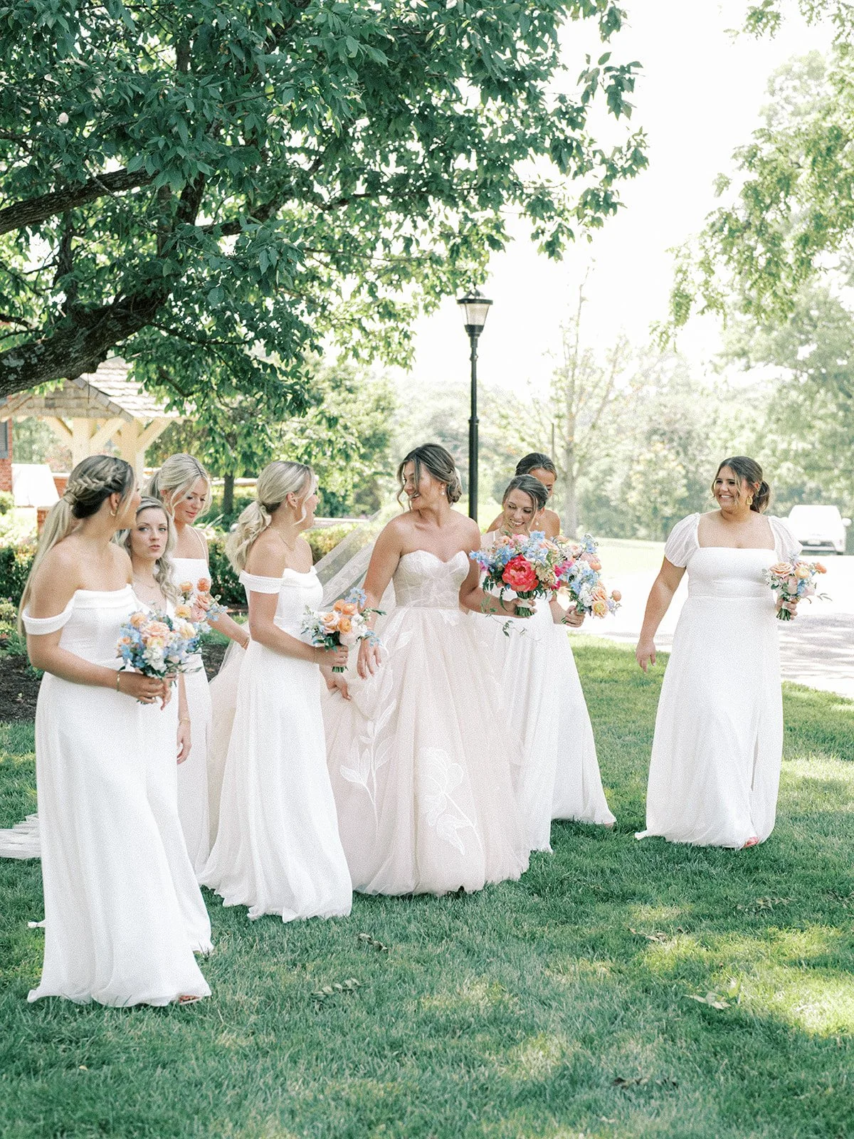 Bride in a white wedding gown with six bridesmaids in white dresses holding bouquets, standing under a large leafy tree on a sunny outdoor lawn in front of the French House Cincinnati