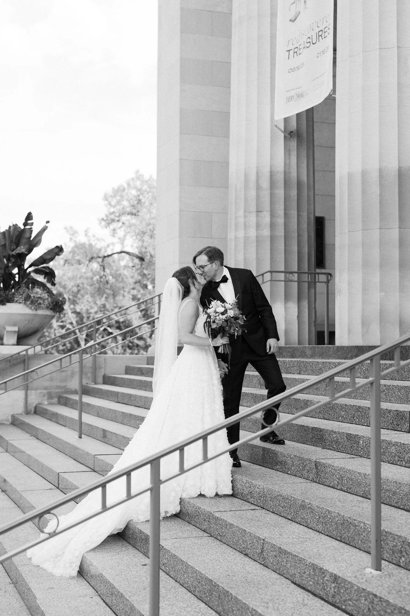 Black and white photo of a bride and groom on the steps of a building, sharing an intimate moment. The bride is in a wedding gown holding a bouquet, and the groom is in a tuxedo leaning in to kiss her.