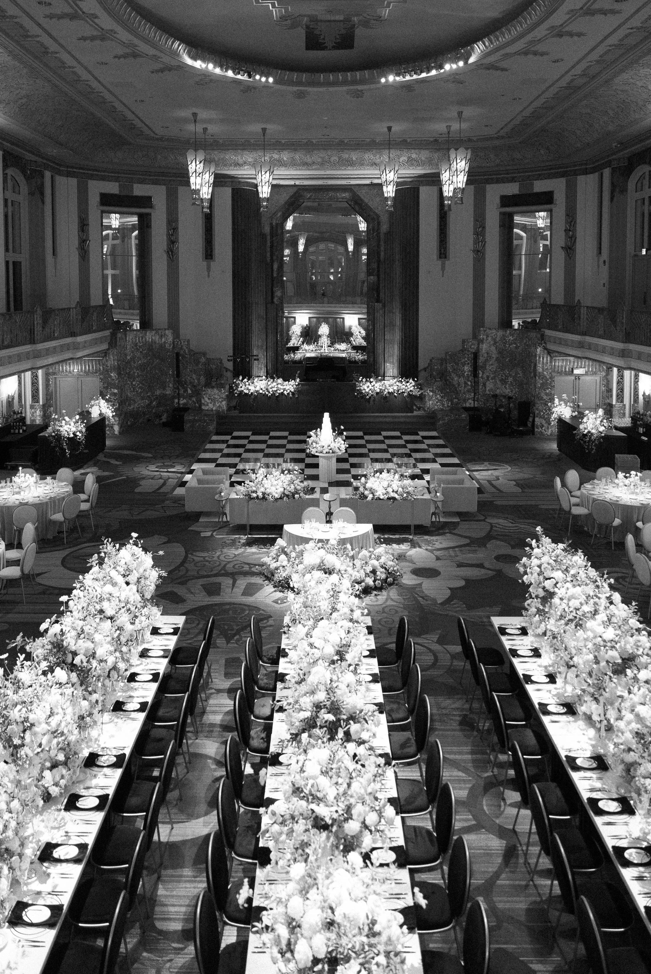 Black and white image of an elegant banquet hall decorated for a formal event, with long tables adorned with floral centerpieces and arrangements, elegant chairs, and a checkered dance floor at the far end.